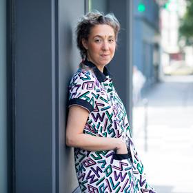 Une personne aux cheveux courts et bouclés est adossée à un mur sombre en extérieur, vêtue d'une chemise à manches courtes ornée d'un motif géométrique audacieux et coloré. L'arrière-plan montre un trottoir et des bâtiments.