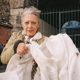 Une femme âgée aux cheveux gris tient un grand tissu blanc drapé. Elle se tient dehors, devant un portail métallique et un mur de pierre, vêtue d'un foulard et d'un manteau clair.