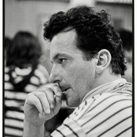 Photographie en noir et blanc d'un homme de profil aux cheveux noirs bouclés, portant une chemise rayée, posant son menton sur sa main et l'air pensif ; arrière-plan flou avec des personnes assises.