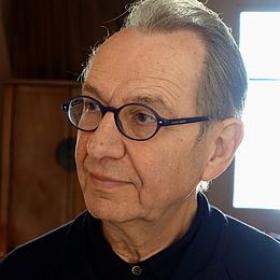 Un homme âgé aux cheveux gris et portant des lunettes regarde de côté. Il porte une chemise sombre et se trouve à l'intérieur, avec la lumière naturelle provenant d'une fenêtre en arrière-plan.