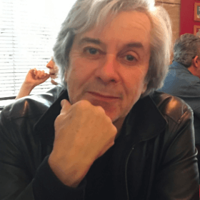 Une personne aux cheveux gris et portant une veste en cuir noir est assise à une table, le menton appuyé sur la main, regardant la caméra. D'autres personnes et une fenêtre avec des stores en arrière-plan.