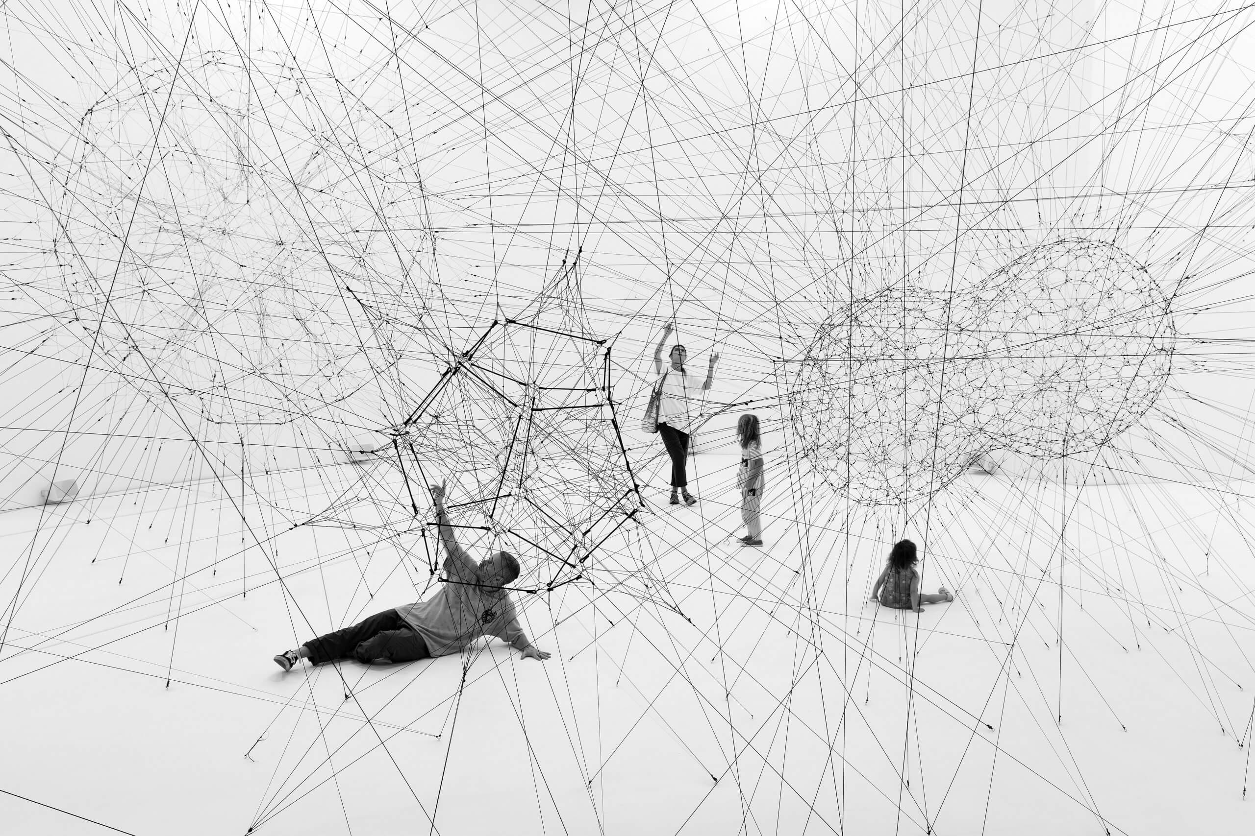 Five people interact with a large geometric art installation made of thin, intersecting wires and polygonal shapes in a white, minimal space. Some people are standing, while others are sitting or lying on the floor.
