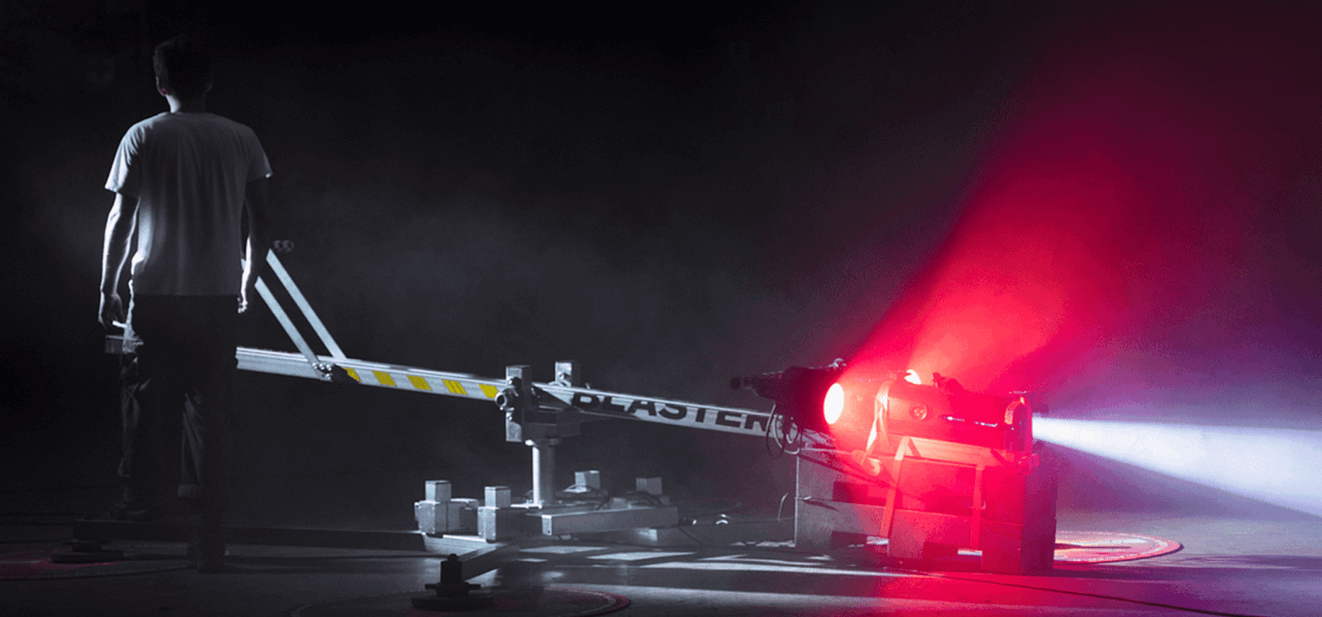 Une personne se tient près d'une grande machine étiquetée « BLASTER », qui émet un puissant faisceau de lumière rouge dans un environnement sombre et enfumé. La scène est éclairée par la lueur rouge de la machine.