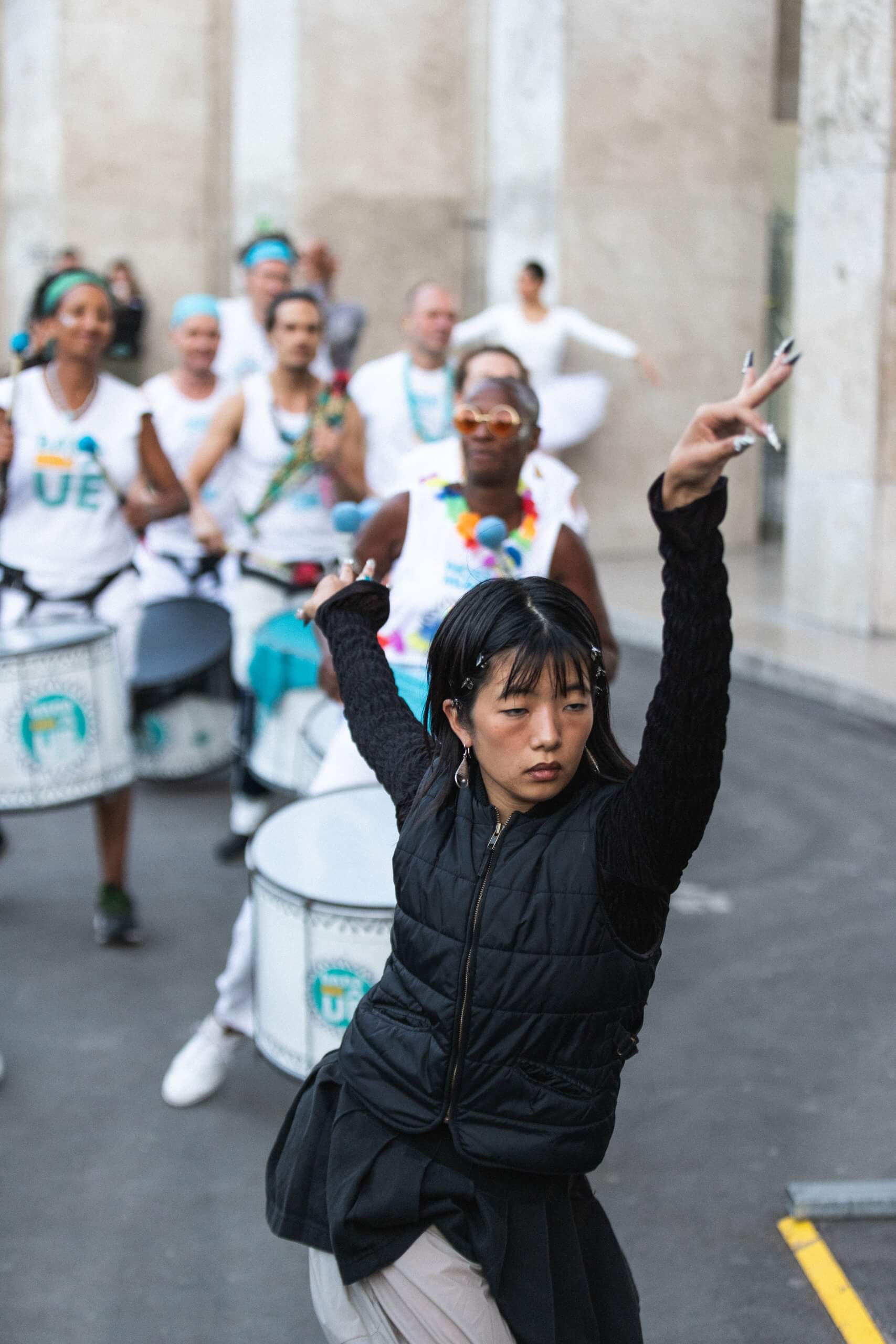 Une femme vêtue d'un gilet noir et de manches sombres danse avec un bras levé devant un groupe de batteurs vêtus de blanc, se produisant en plein air dans une rue de la ville.
