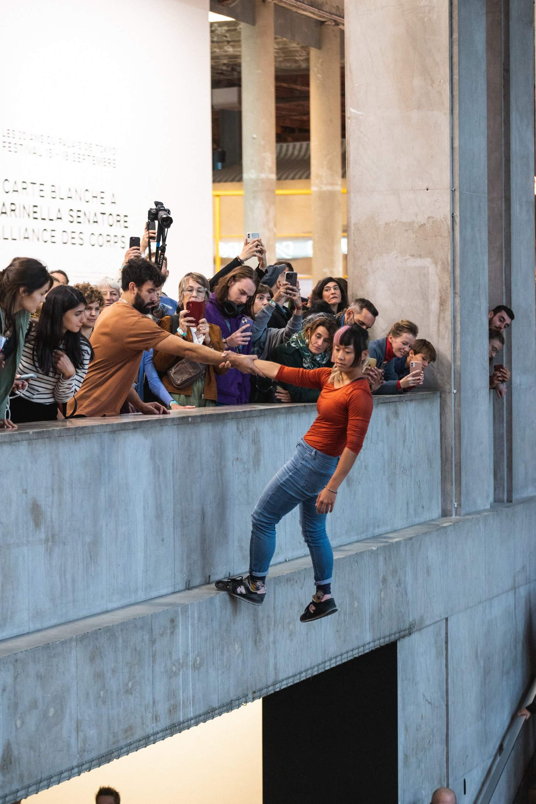 Une femme se tient en équilibre précaire sur une étroite corniche au-dessus d'un précipice, tandis qu'un groupe de personnes, sur un balcon, tendent leurs mains et leurs téléphones, certains aidant ou prenant des photos. Le décor semble se dérouler à l'intérieur d'un immeuble moderne.