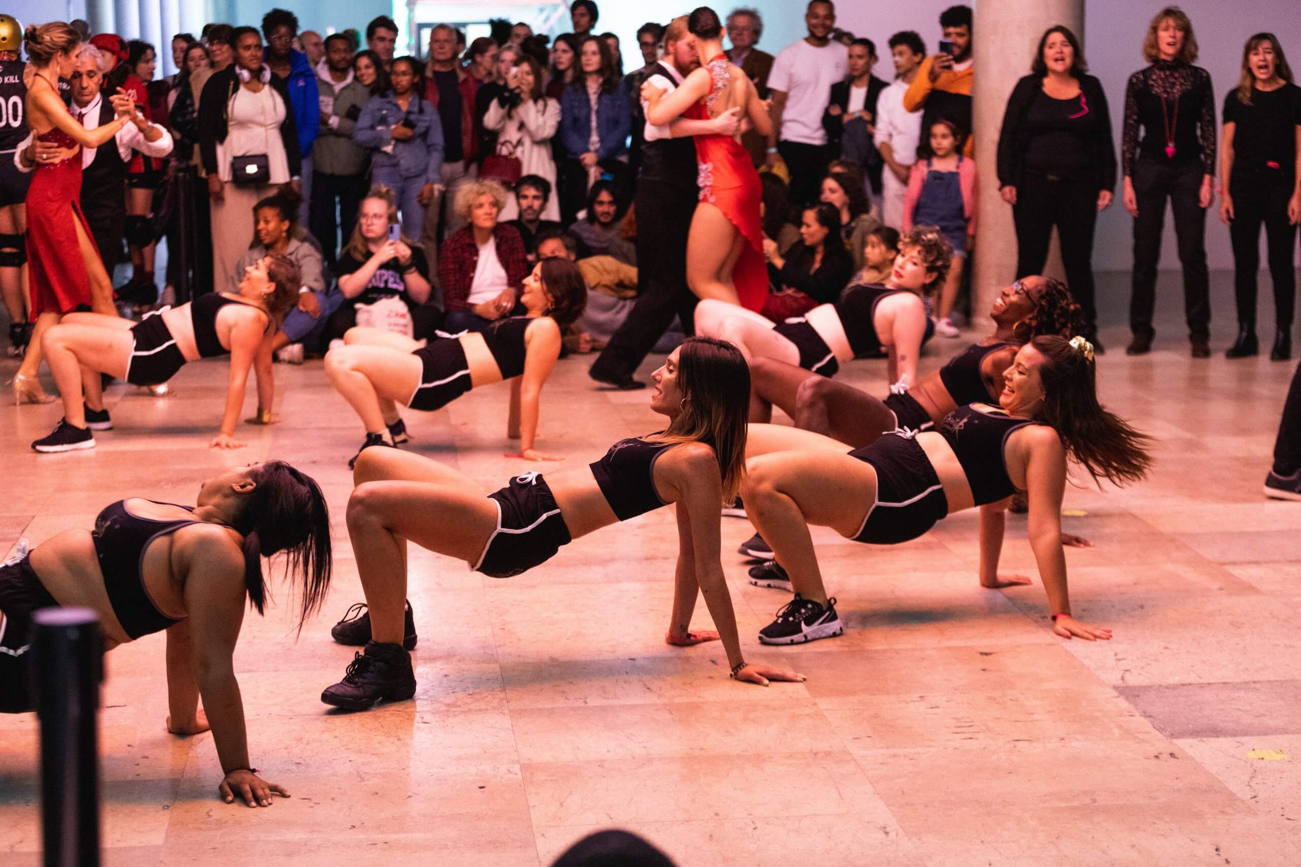 Un groupe de danseurs en tenue de sport noire exécute une chorégraphie synchronisée au sol devant un large public en salle. Les spectateurs observent attentivement, certains debout, d'autres assis ou prenant des photos.