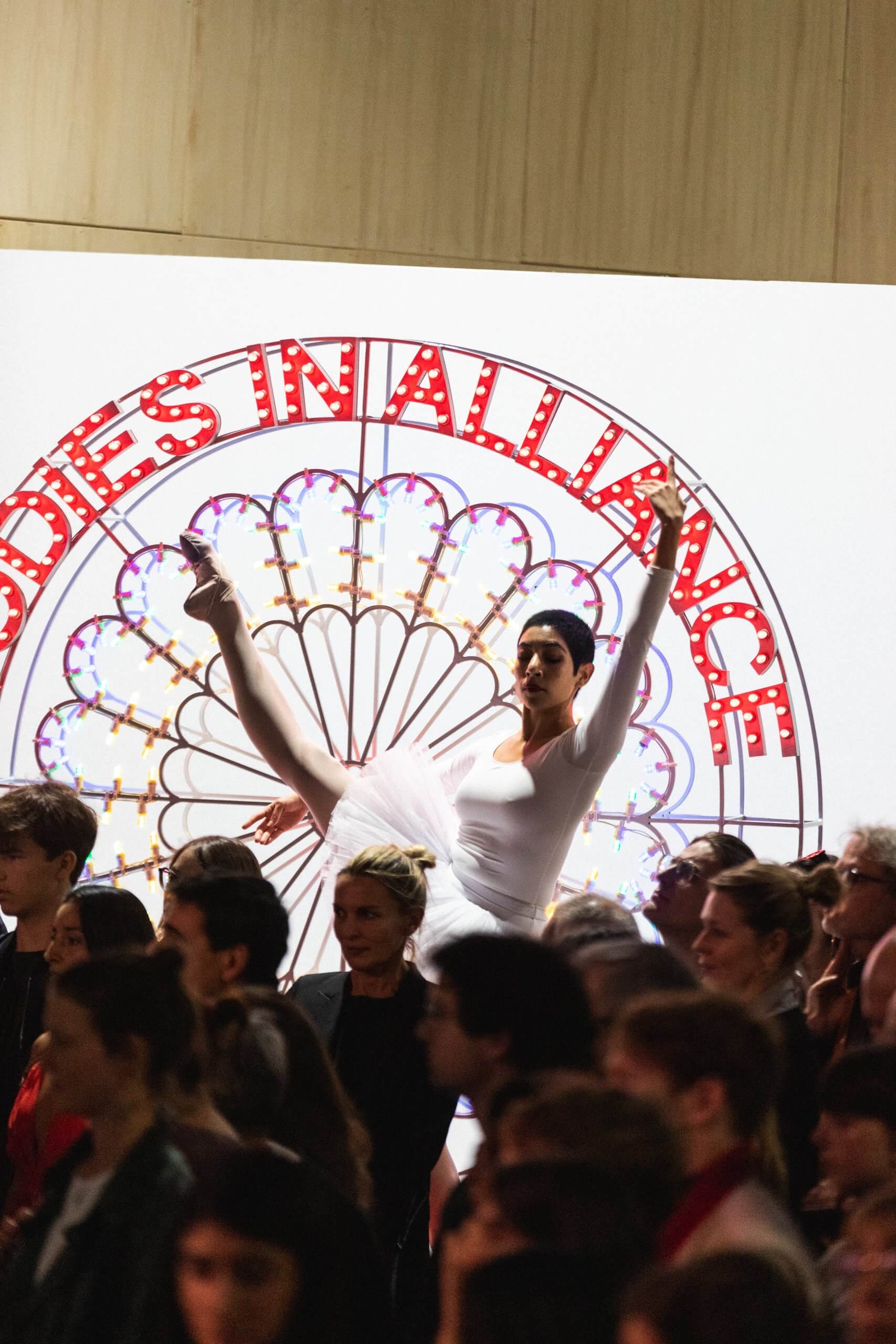 Un danseur de ballet en costume blanc exécute une arabesque sur une jambe devant un grand panneau indiquant CORPS EN ALLIANCE, avec une foule de personnes debout au premier plan.