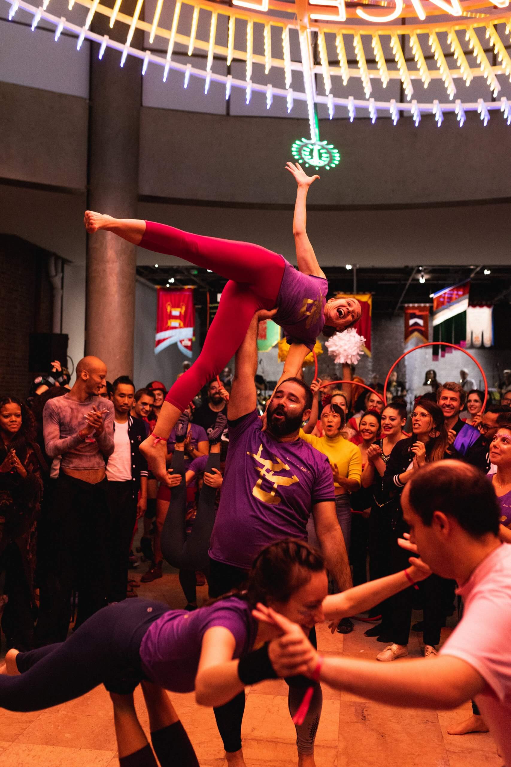 Un homme en chemise violette soulève une femme souriante en leggings rouges au-dessus de sa tête dans un espace intérieur bondé, tandis que d'autres se produisent et qu'un groupe de personnes debout à proximité regardent et applaudissent.
