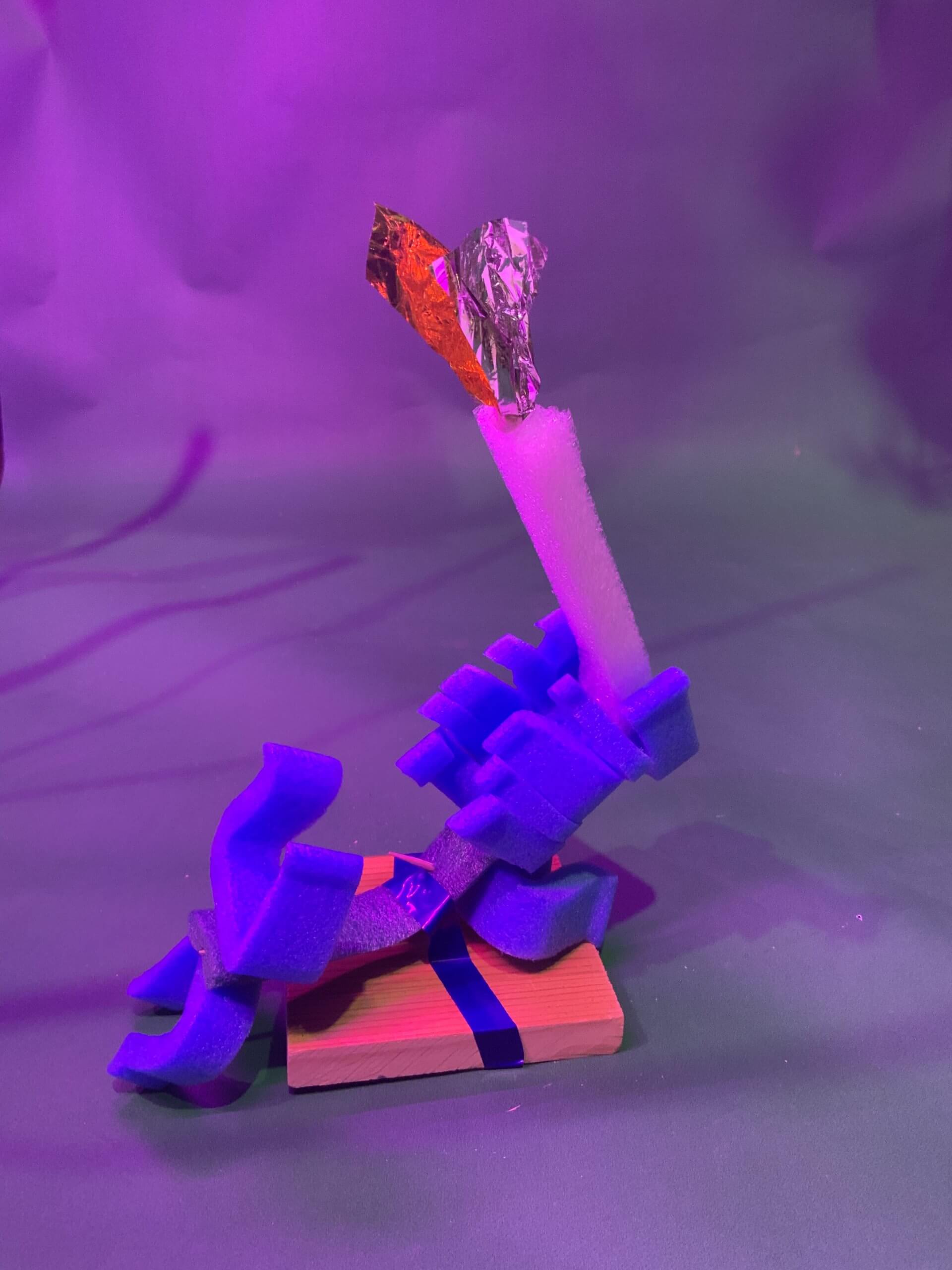 Une sculpture composée de pièces de mousse bleue et blanche disposées sur une base en bois, avec un morceau de feuille d'argent et d'orange attaché au sommet, sur un fond éclairé par un violet.