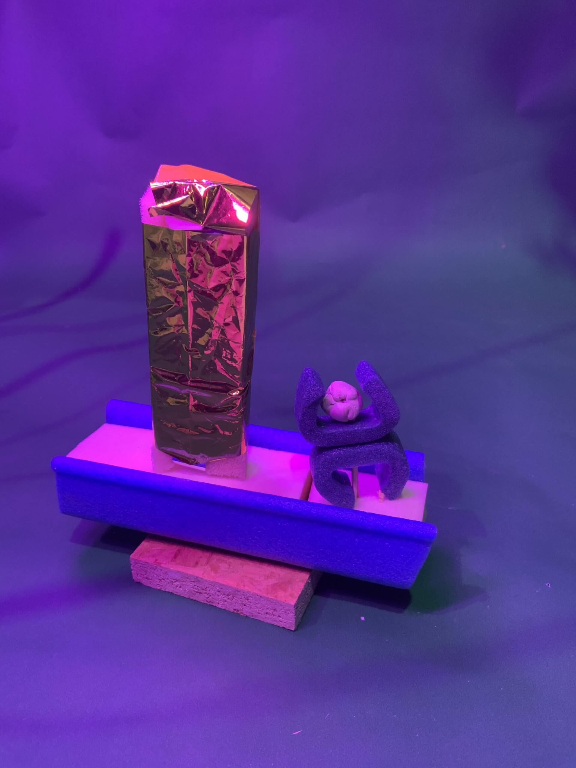 Une petite sculpture représentant un grand objet rectangulaire doré et une figure abstraite violette à tête ronde, tous deux posés sur une plate-forme faite de divers matériaux, sous un éclairage violet.