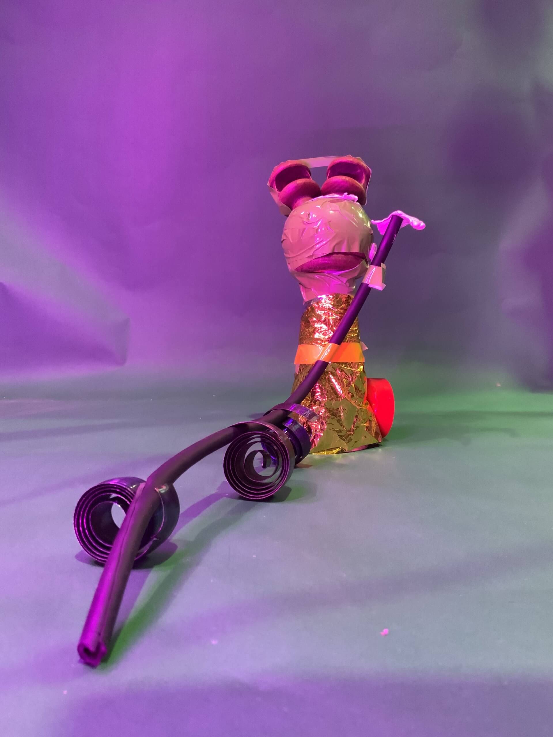 Une petite sculpture verticale fabriquée à partir de matériaux ménagers, notamment de feuilles d'or, de ruban adhésif et de tubes en plastique, se dresse sur une surface verte avec un câble noir enroulé au premier plan sur un fond violet.