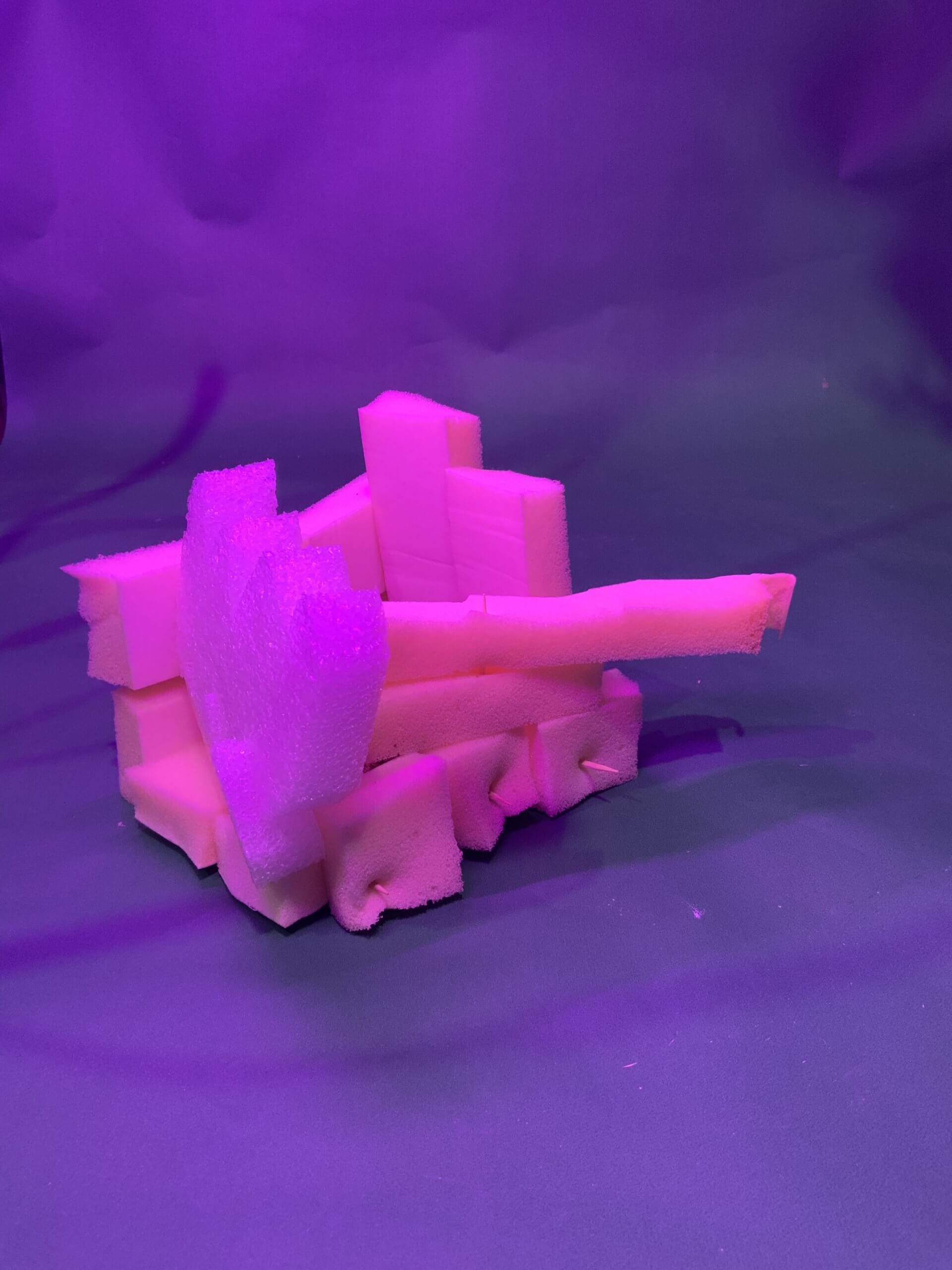 Plusieurs morceaux de mousse rose sont empilés et disposés pour ressembler à un réservoir, éclairé par un éclairage violet, sur un fond sombre uni.