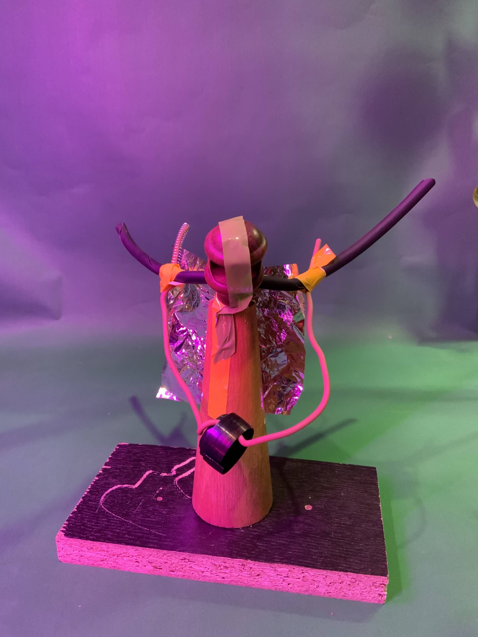 Un cône en bois repose debout sur une base rectangulaire. Il est doté d'ailes en aluminium, de cure-pipes et de fils métalliques fixés avec du ruban adhésif, ainsi que d'un objet cylindrique noir suspendu près de la base. L'arrière-plan est éclairé par des lumières violettes et vertes.