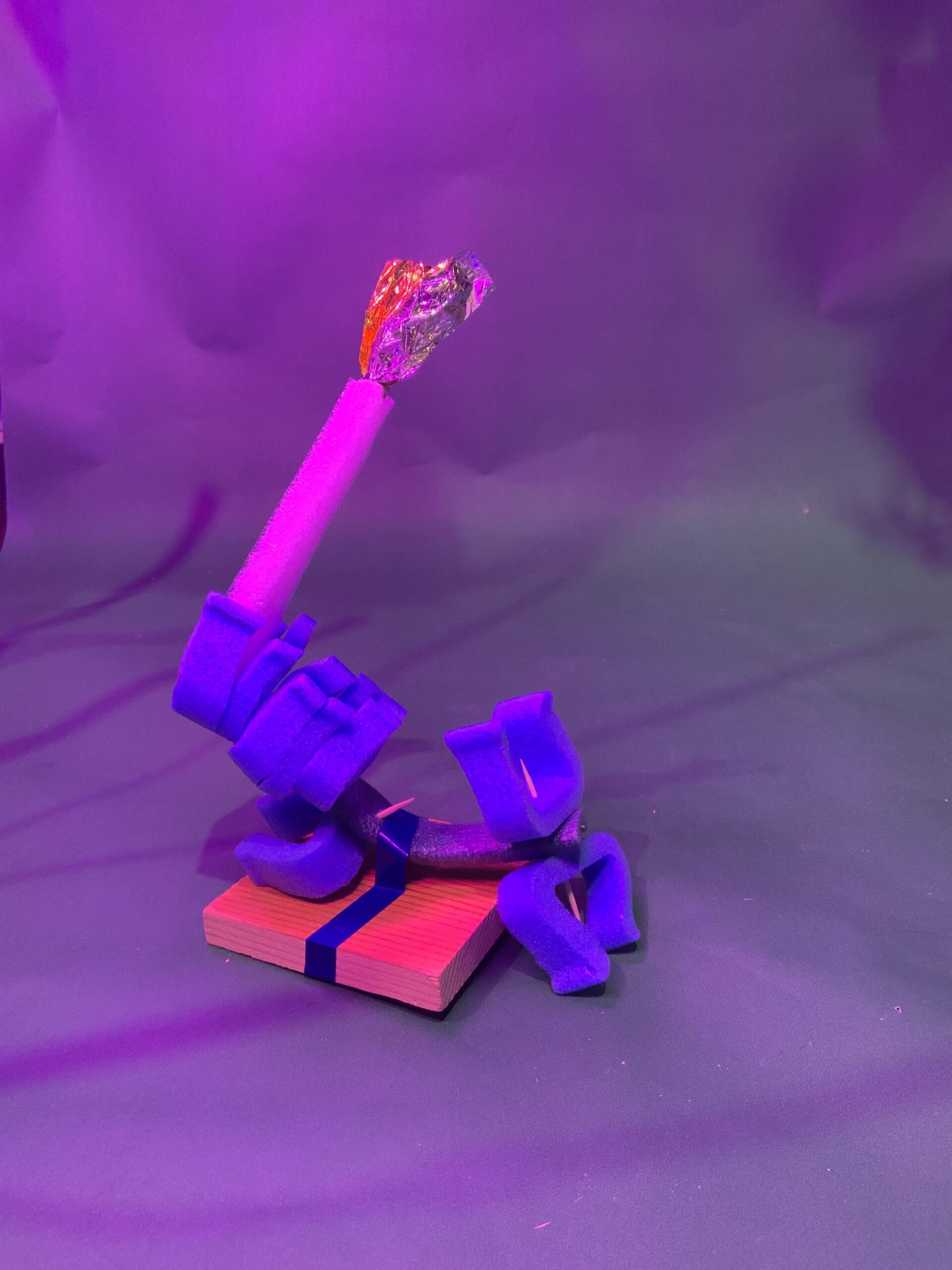 Un objet sculptural composé de mousse violette, de bois et de matériaux métalliques est présenté sur un socle en bois sur un fond violet éclairé. L'objet présente plusieurs formes courbes et rectangulaires.