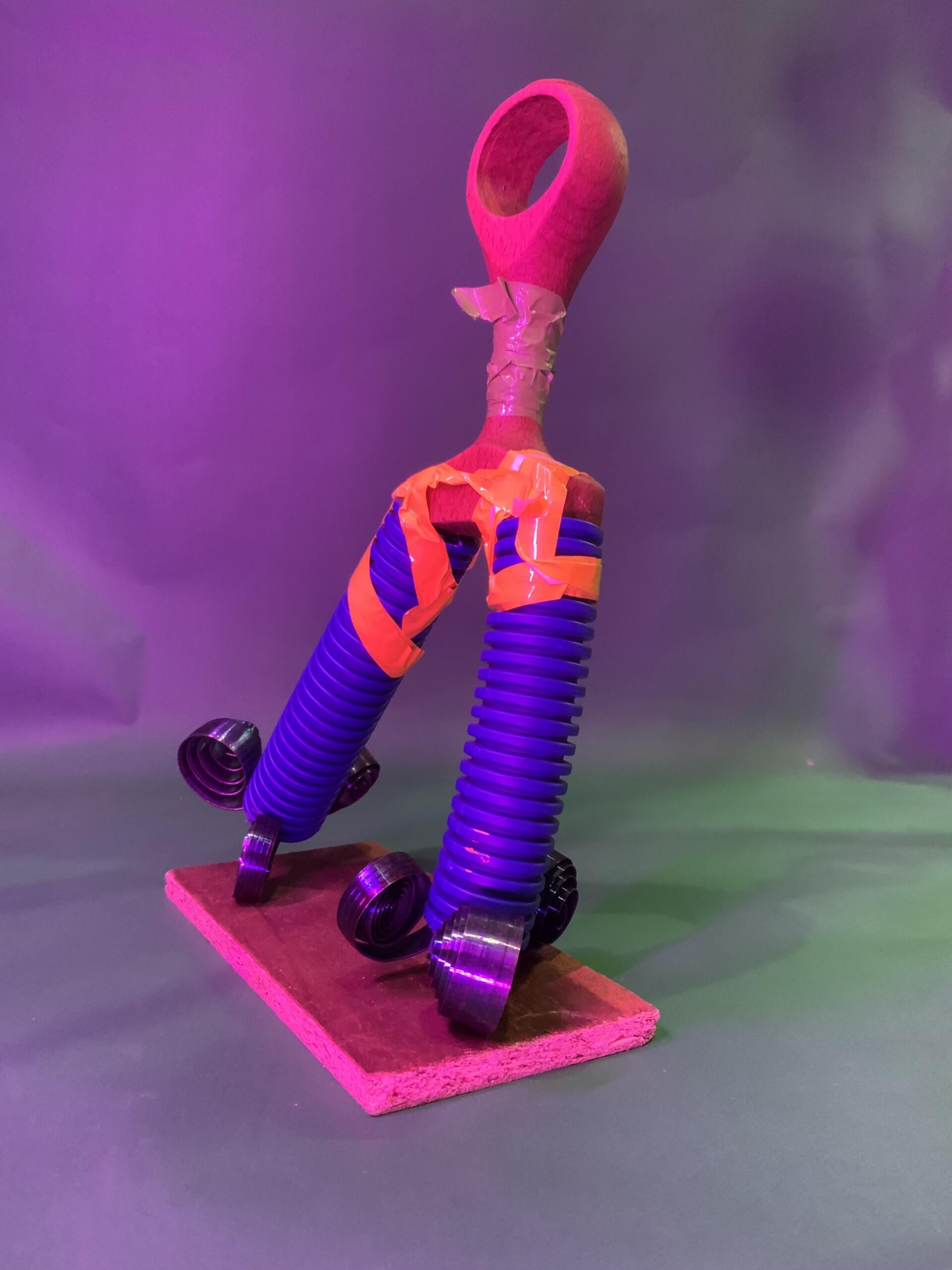 Une sculpture composée d'une cuillère en bois, de ruban adhésif orange, de tubes ondulés violets et de pièces métalliques enroulées repose sur une base rectangulaire en bois, sur un fond violet avec un éclairage dramatique.
