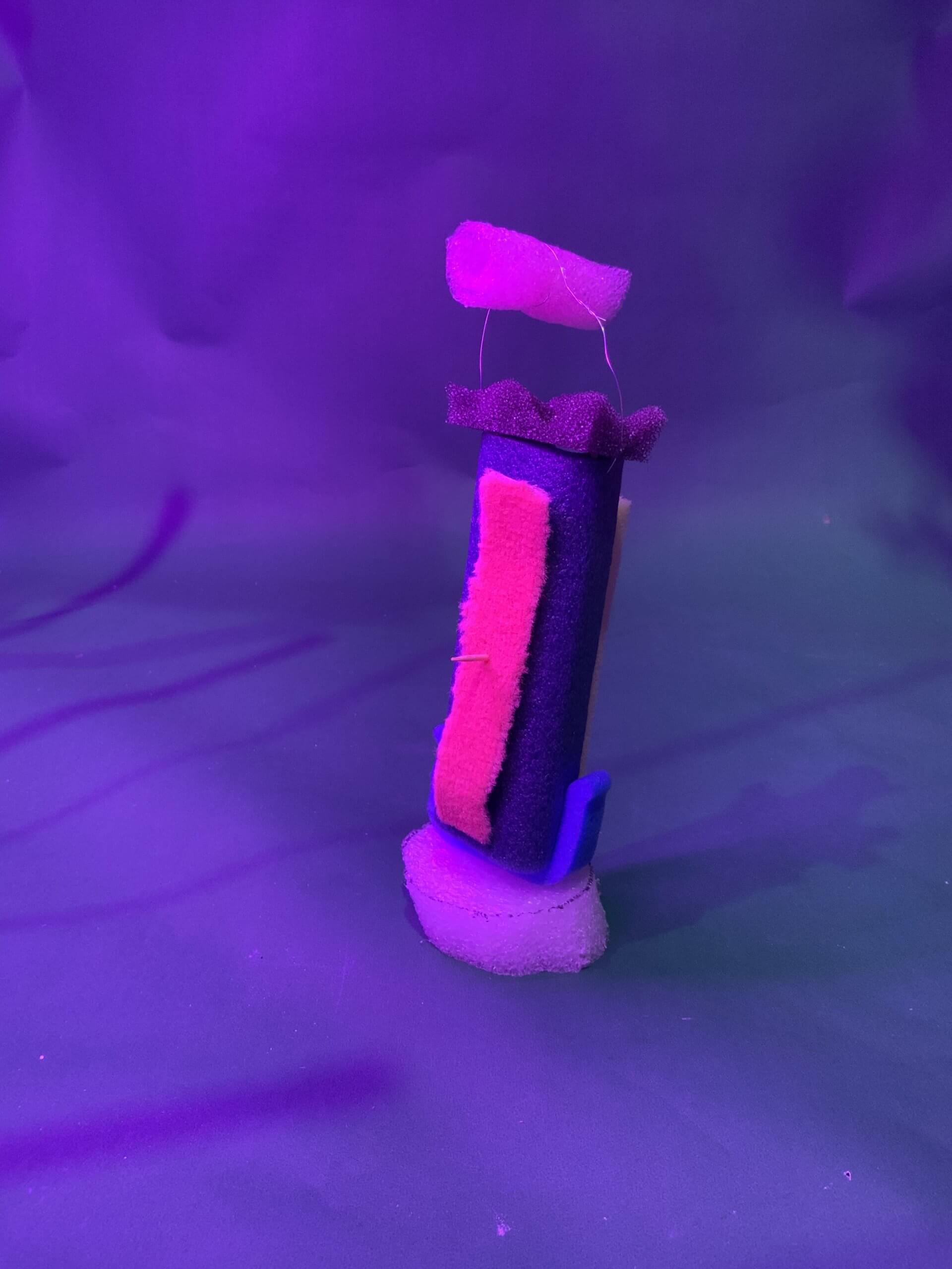 Une sculpture abstraite verticale réalisée à partir d'éponges de différentes couleurs repose sur une base ronde en éponge, éclairée par un éclairage violet sur un fond violet assorti.