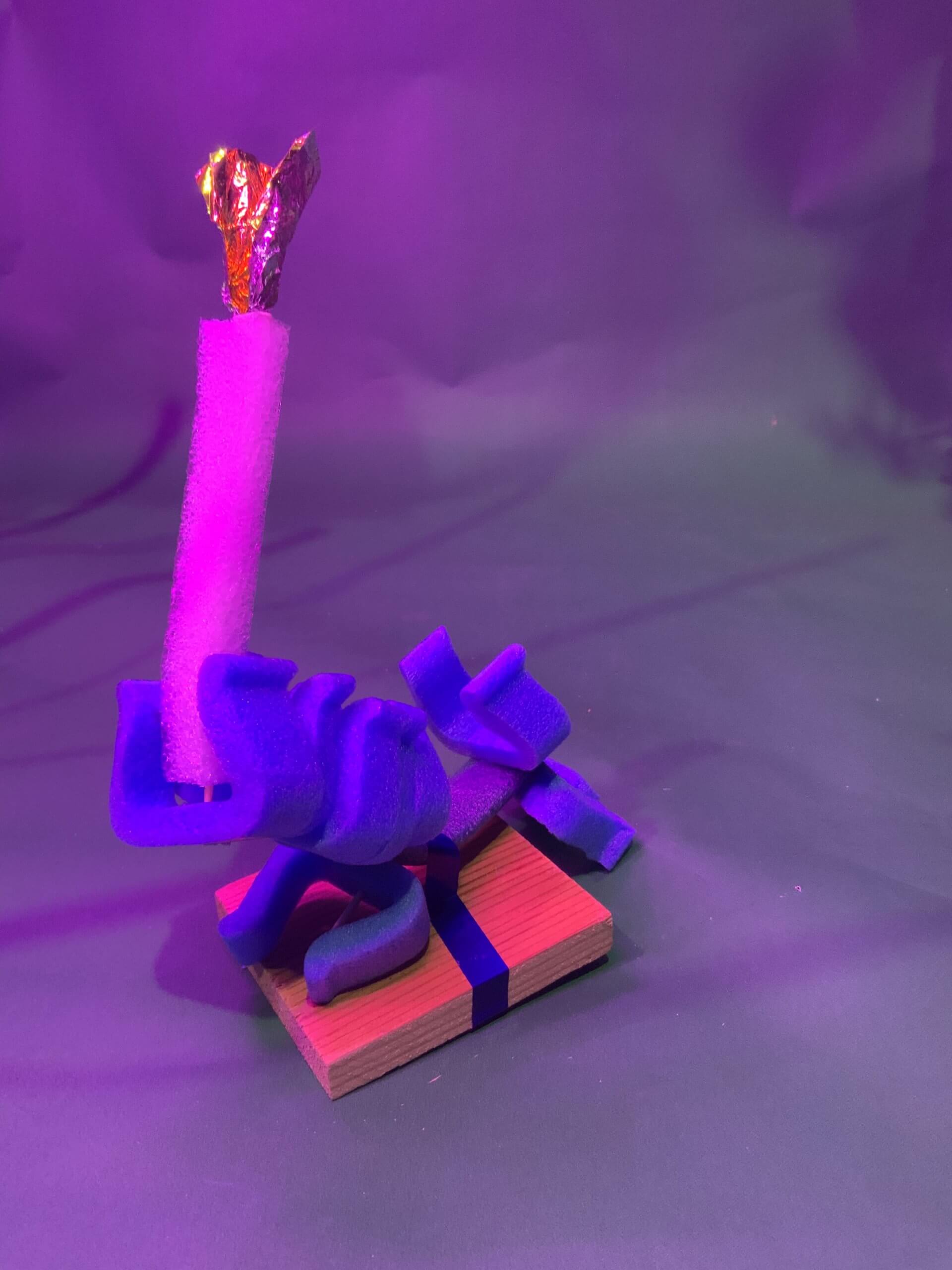 Une petite sculpture abstraite repose sur une base en bois, comportant des formes en mousse bleue, une pièce cylindrique blanche et un élément métallique doré au sommet, le tout sur un fond éclairé en violet.