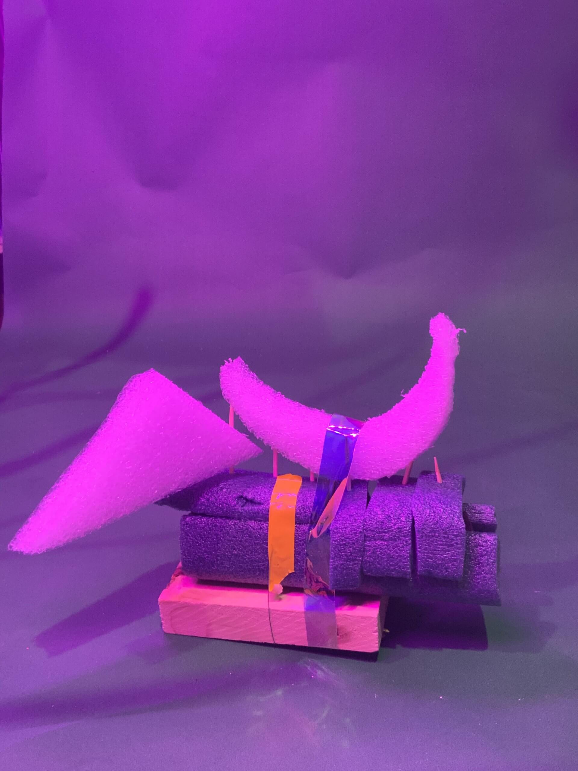 Une sculpture composée de pièces de mousse violette, de ruban adhésif orange et violet et d'un bloc de bois est présentée sur un fond violet sous un éclairage violet. Les pièces de mousse sont empilées et disposées selon des formes géométriques.