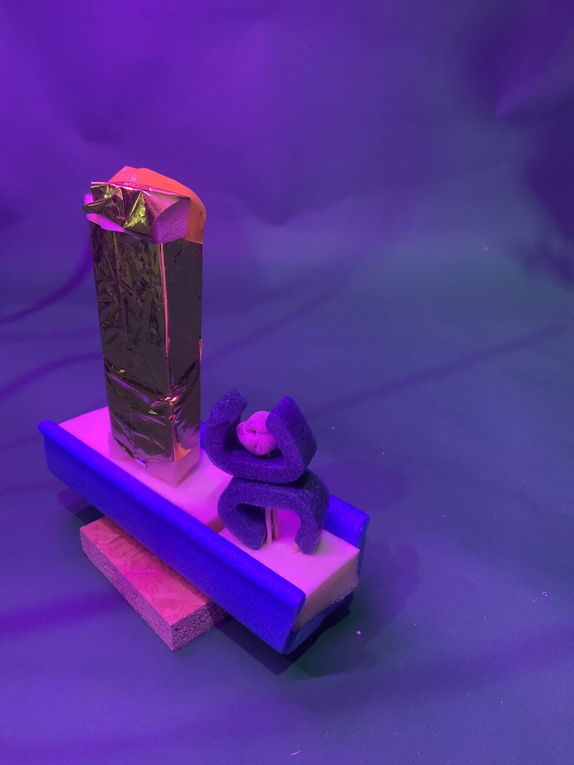 Une petite sculpture abstraite avec un bloc rectangulaire enveloppé dans une feuille d'or debout à côté d'une éponge bleue et de formes en mousse violette, le tout placé sur une base en mousse avec des morceaux de bois en dessous, sur un fond violet.