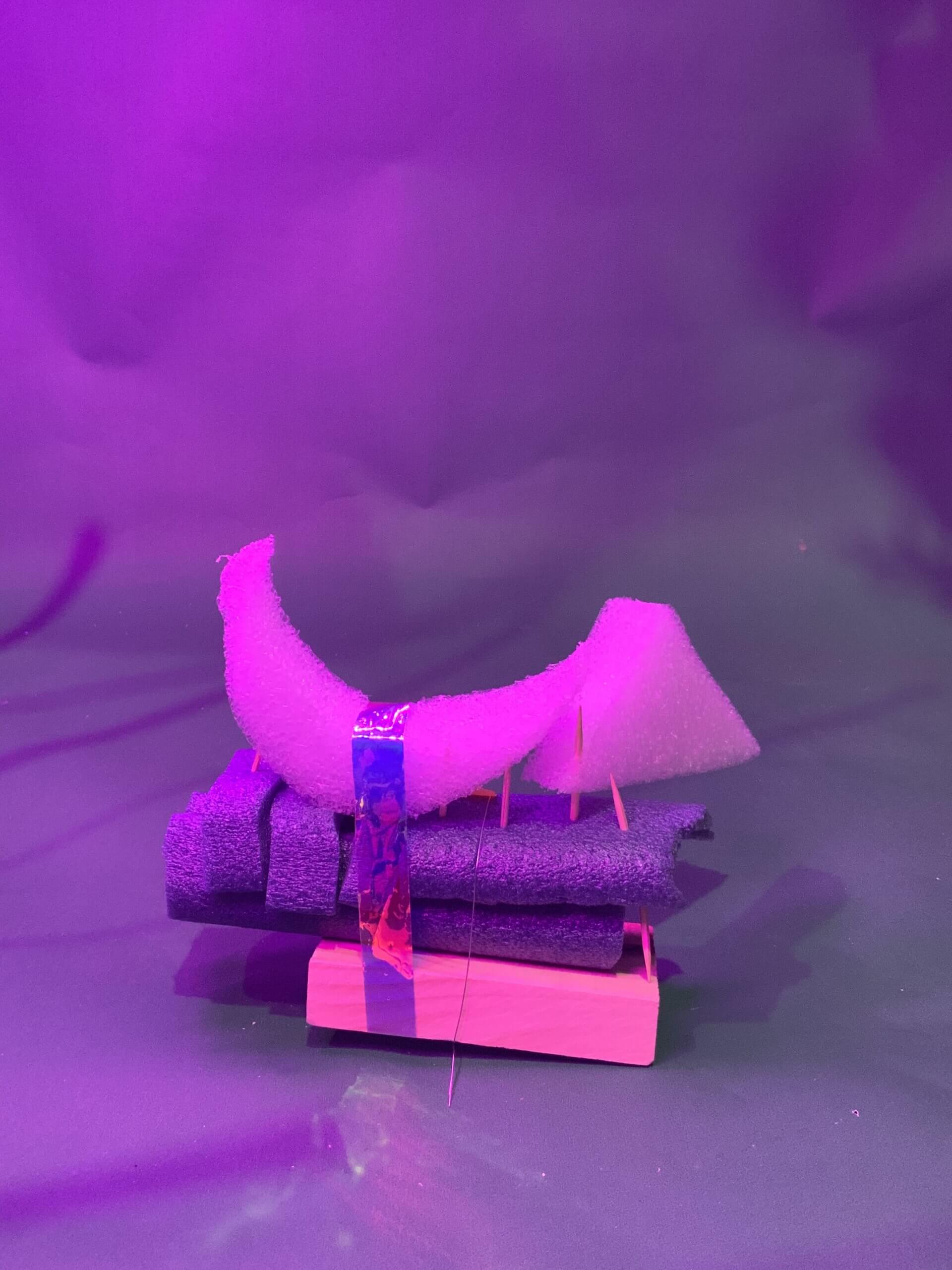 Un morceau incurvé de mousse blanche est soutenu par des cure-dents et du ruban adhésif sur des serviettes violettes empilées et un bloc de bois rectangulaire, le tout sur un fond violet.