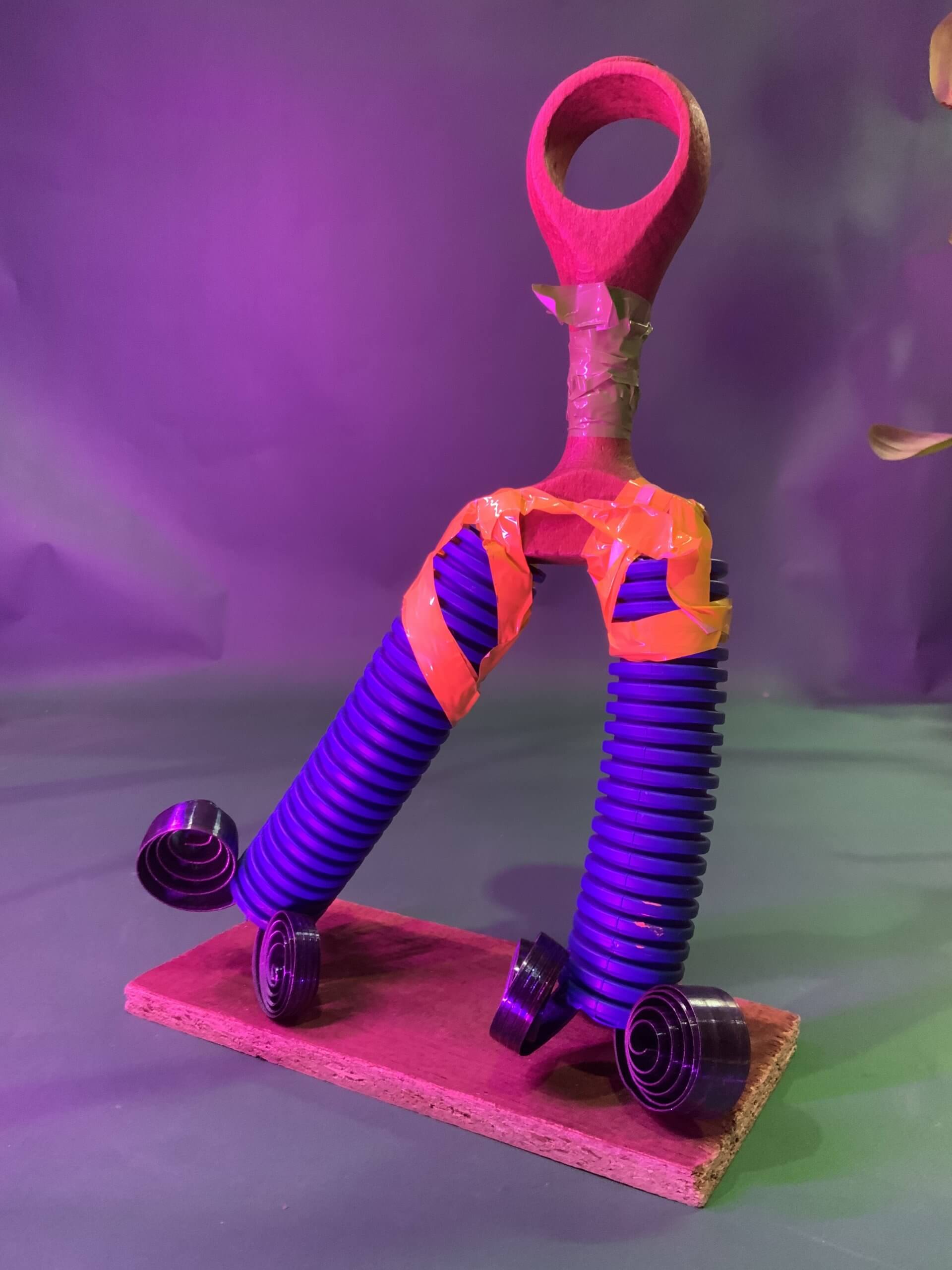 Une sculpture réalisée à partir de deux ressorts bleus enroulés fixés avec du ruban adhésif orange sur un manche en bois, formant une figure debout sur une base rectangulaire en bois, le tout sur un fond violet.