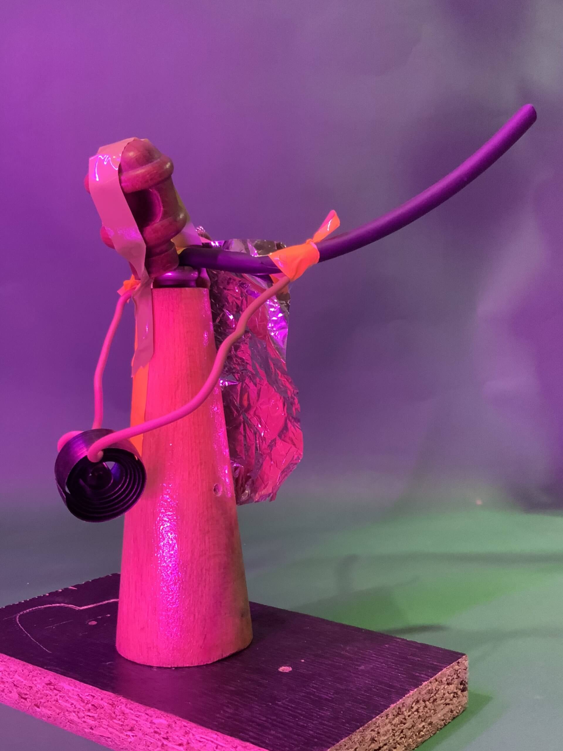 Une sculpture composée d'un cône en bois, d'un marteau, de fil de fer, de papier d'aluminium, de ruban adhésif orange et d'une bobine noire, assemblés sur une base rectangulaire en bois, photographiée sous un éclairage rose et vert.