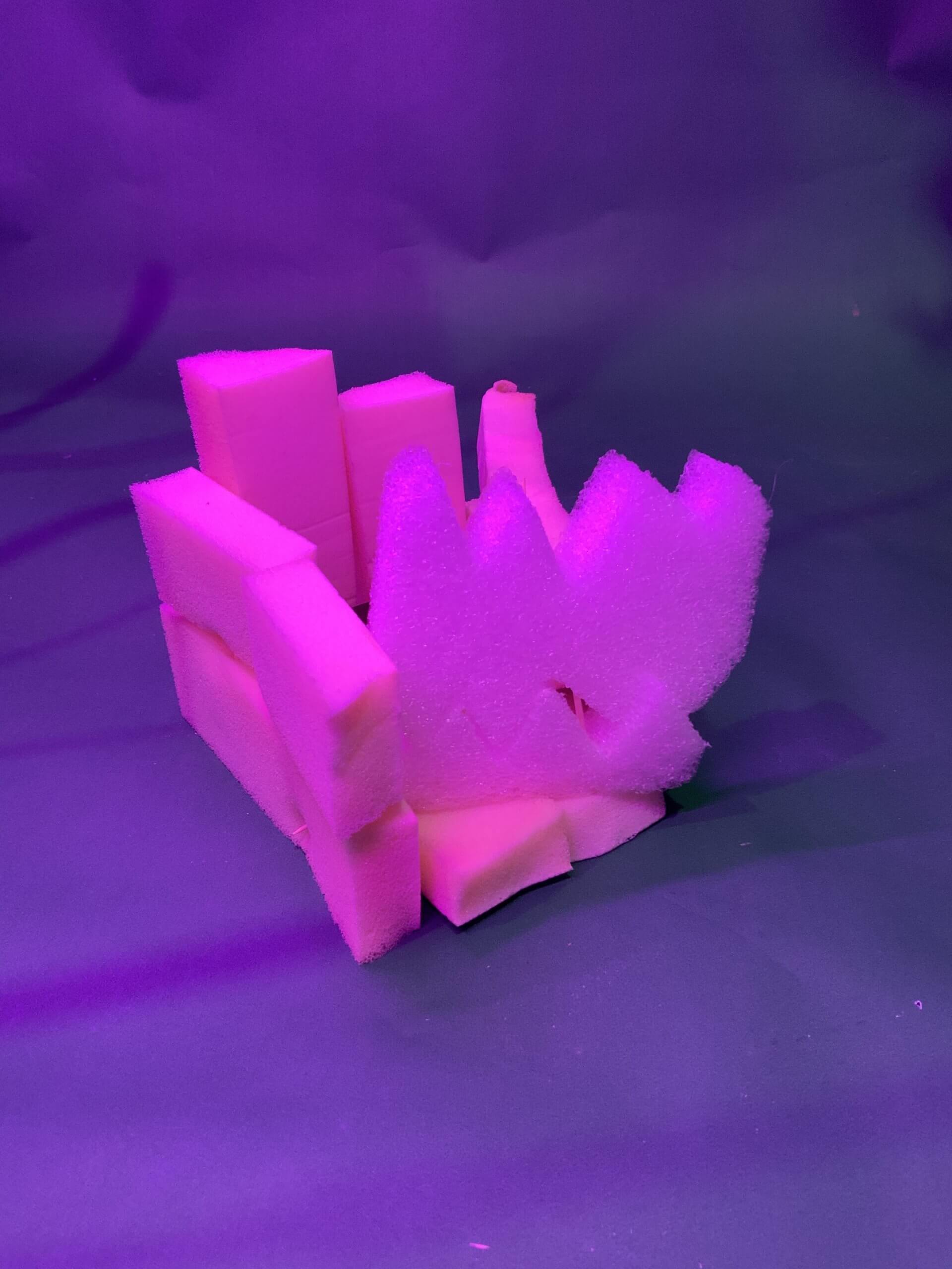 Une petite sculpture abstraite composée de morceaux de mousse rose et blanche disposés verticalement et en angle, sur un fond violet avec un éclairage dramatique.