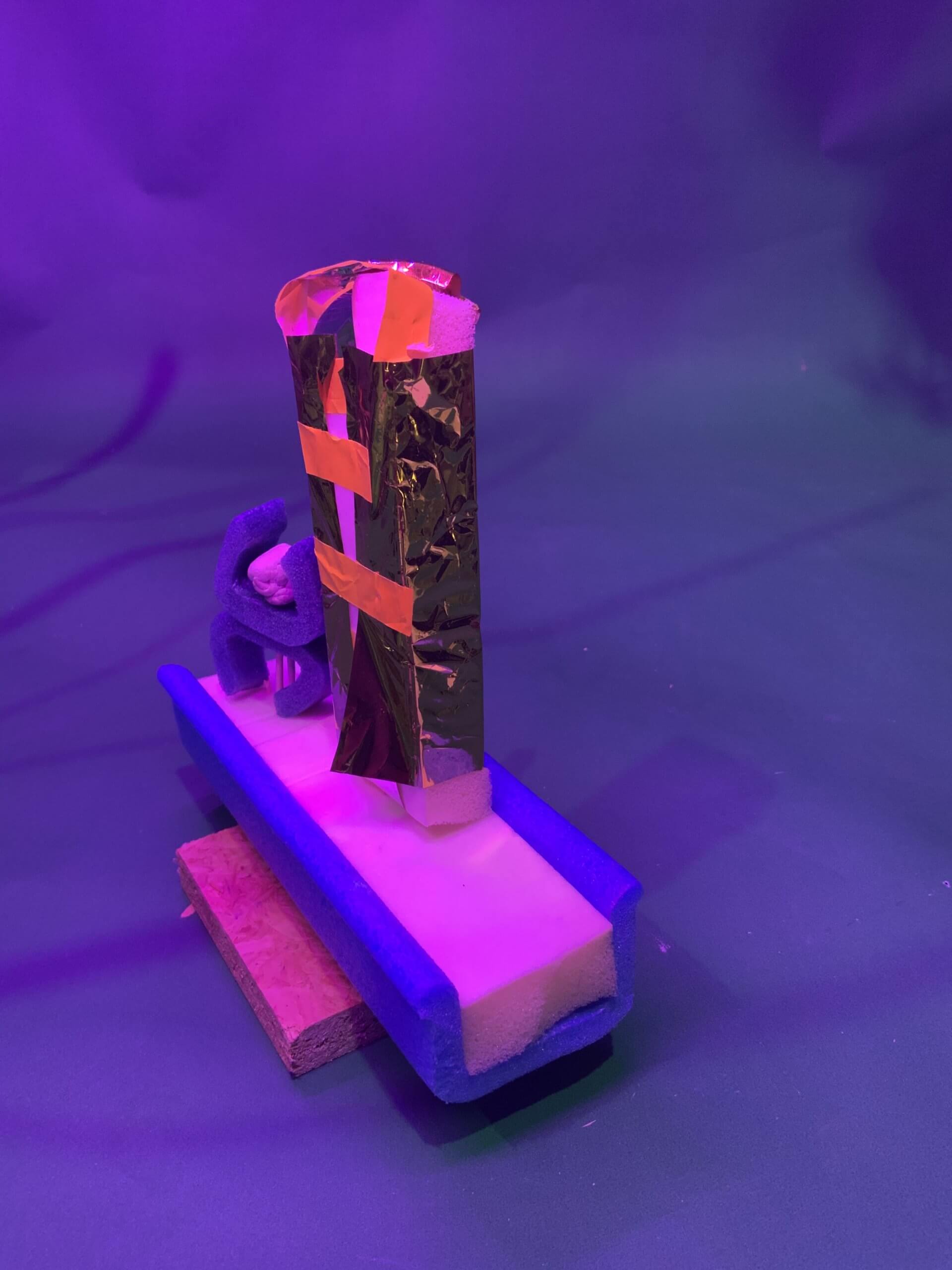 Une maquette artisanale, réalisée en mousse, éponge, feuille d'or et ruban adhésif orange, se détache sur un fond violet et vert sous un éclairage violet. La structure ressemble à une petite sculpture abstraite.