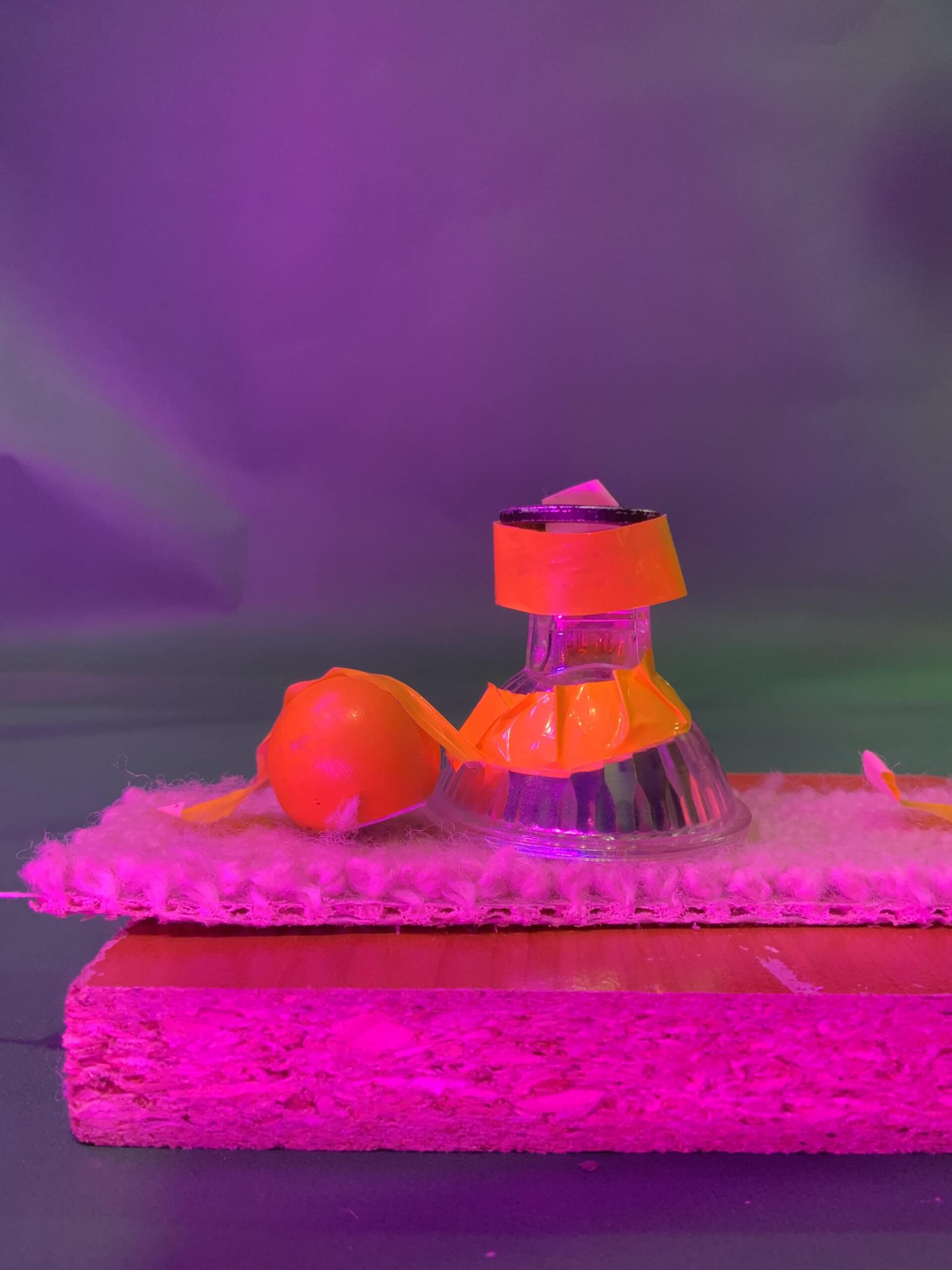 Une composition de nature morte comprenant une boule orange et un verre à l'envers avec du ruban adhésif orange dessus, placé sur un tapis blanc texturé au sommet d'un bloc de bois rose, avec un fond violet.