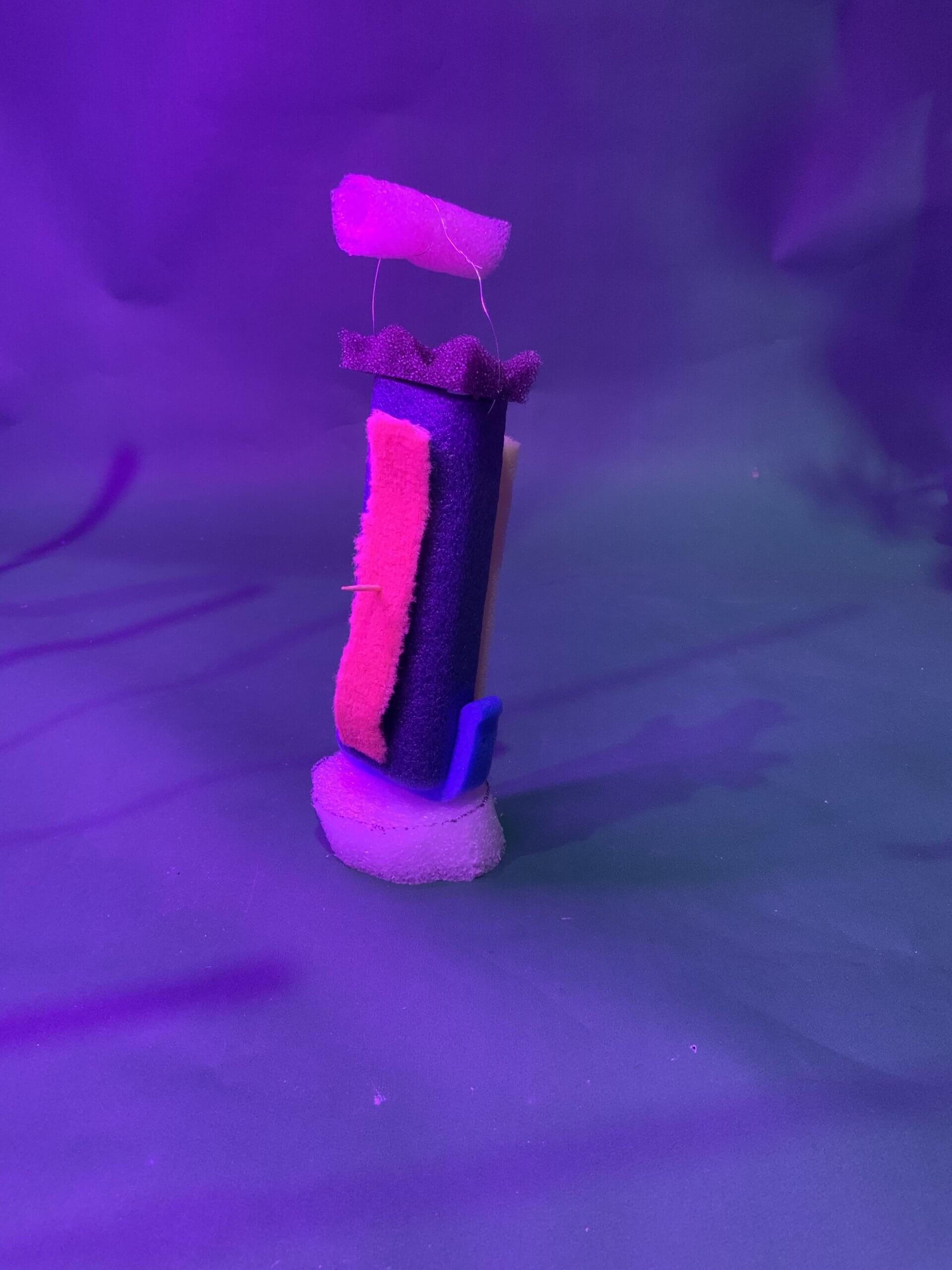 Une petite sculpture abstraite composée d'éponges colorées et de morceaux de mousse est exposée verticalement sur une surface verte éclairée par un effet violet. La structure comprend des éléments en éponge rose, bleu, noir et blanc.