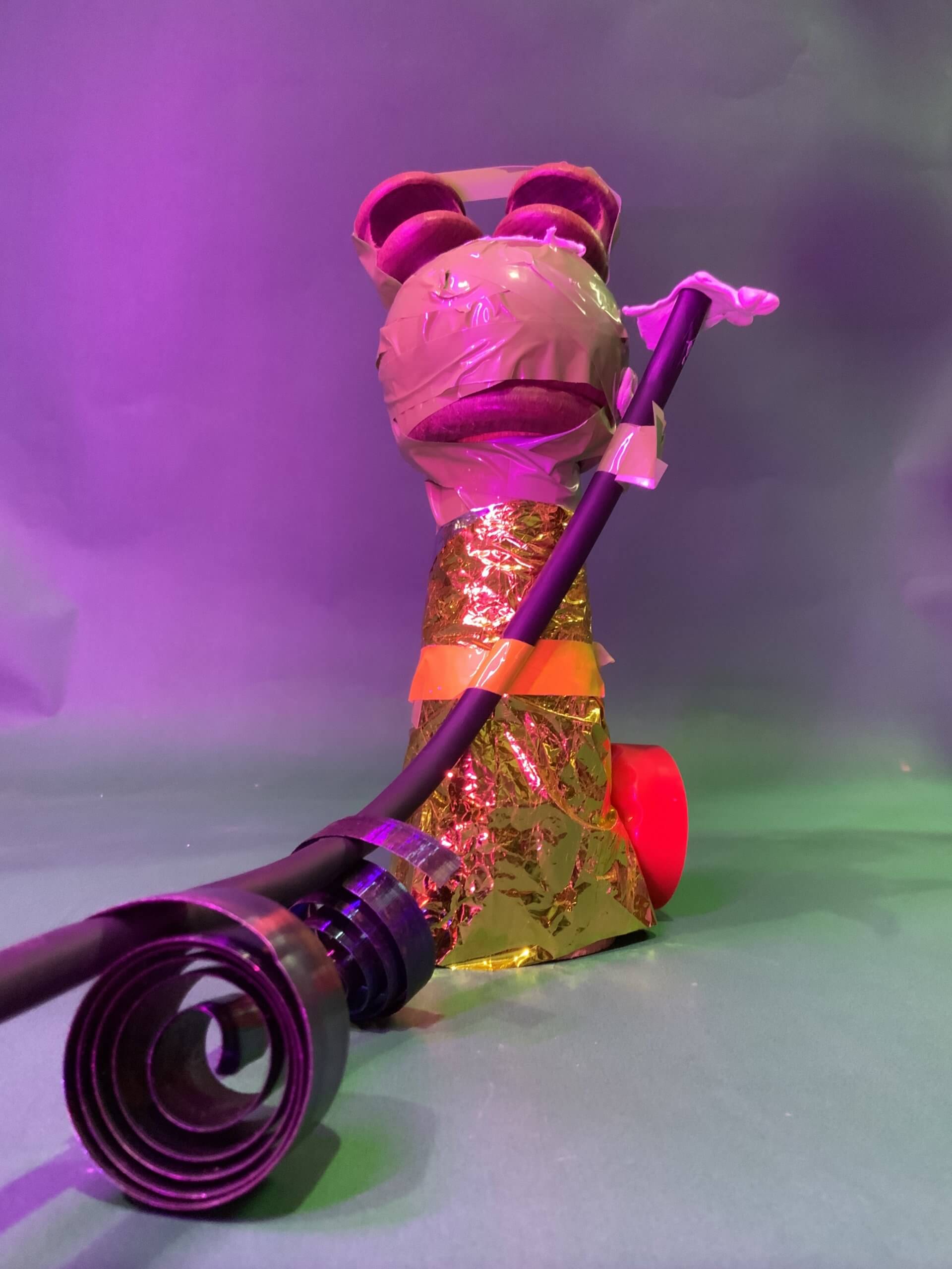 Une sculpture de créature faite maison, réalisée à partir d'articles ménagers, notamment de feuilles d'or, de ruban adhésif, de pièces en plastique rouge et de tubes foncés, est posée sur une surface verte sous un éclairage violet.