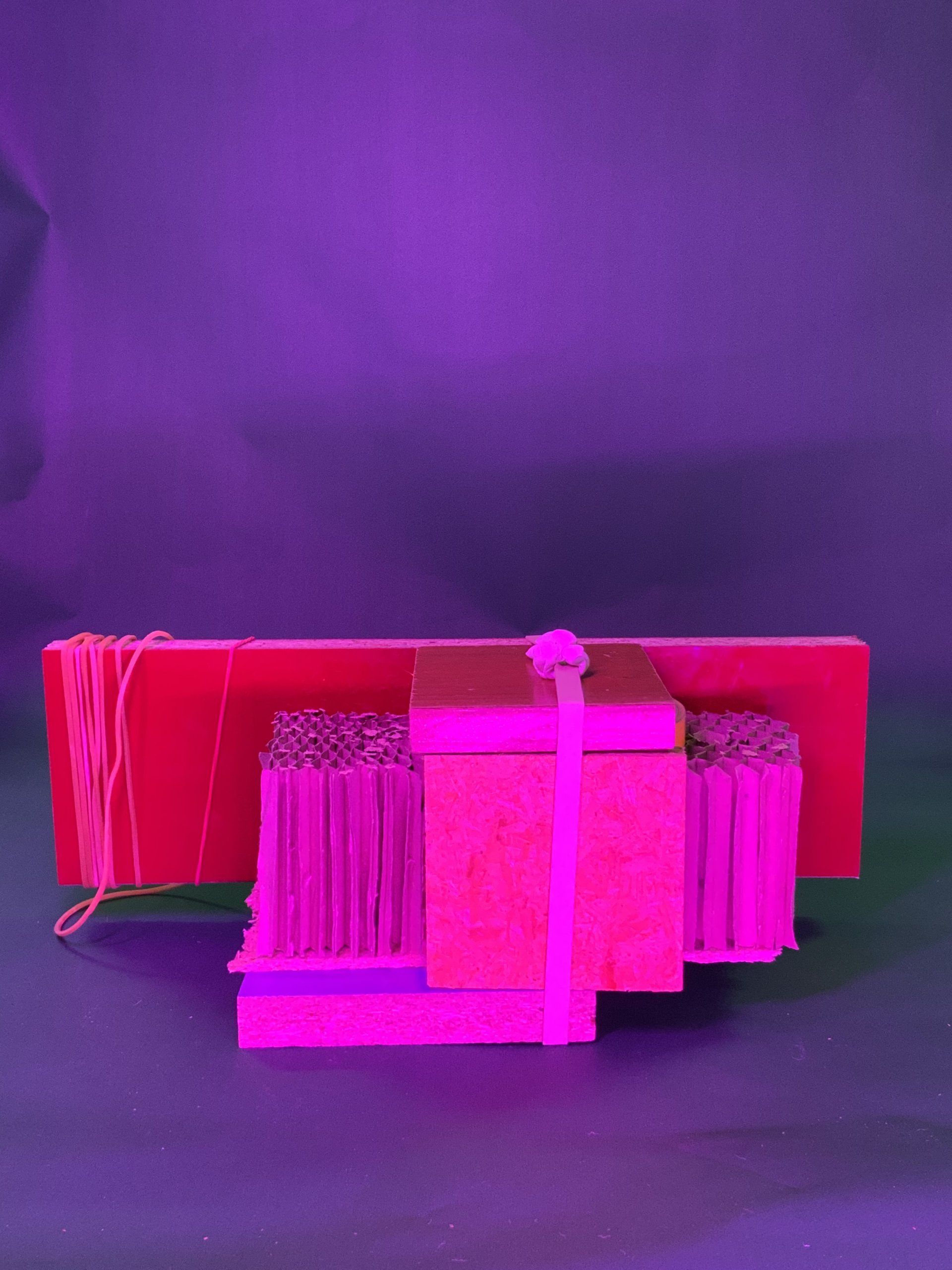 Une pile de pièces rectangulaires en carton est reliée par des élastiques et disposée sur un fond violet. Les objets sont illuminés par un éclairage rose et violet.