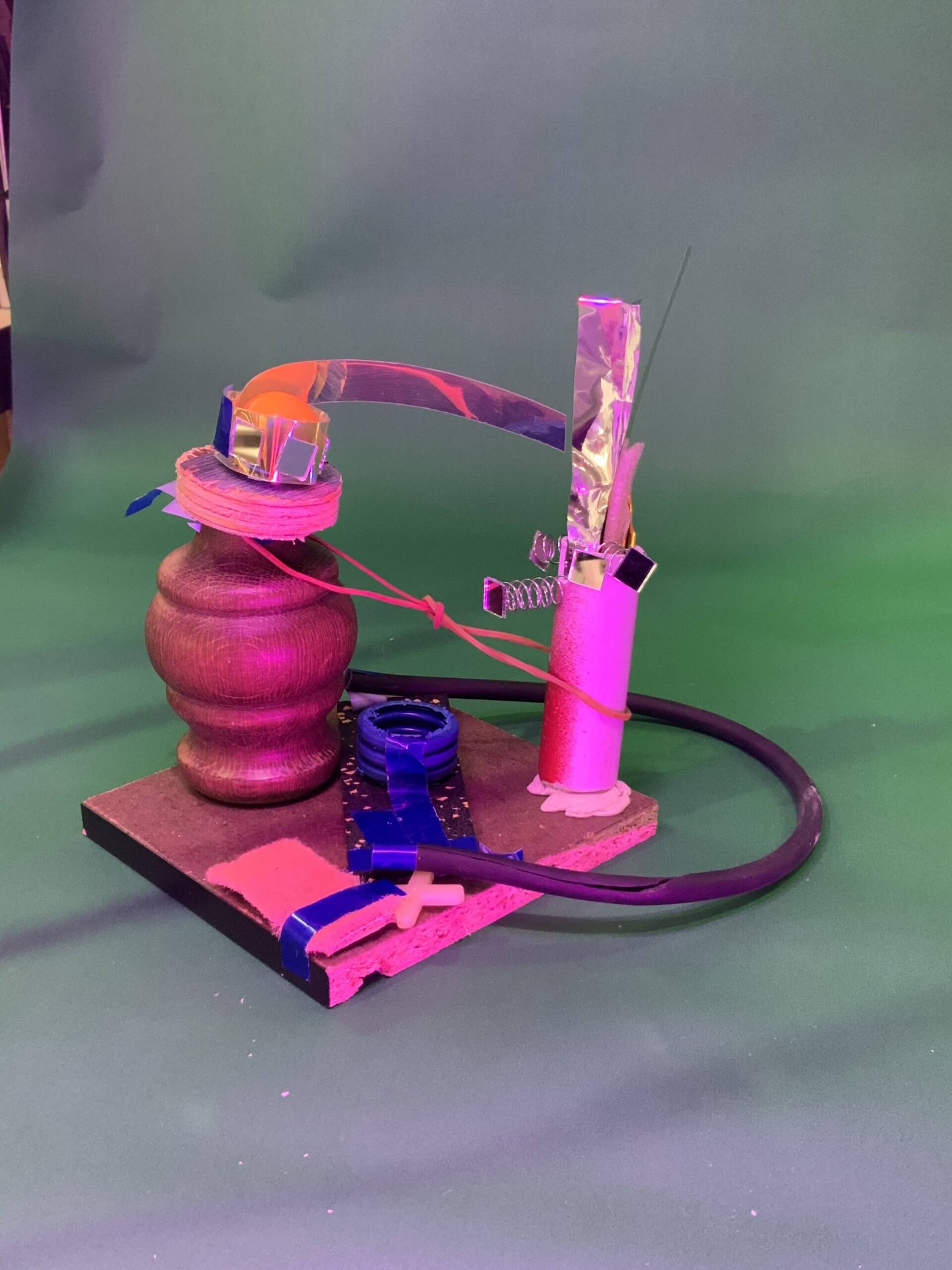 Une petite sculpture abstraite en bois, métal, plastique et carton repose sur un fond vert. L'ensemble comprend un axe en bois, des fils, du papier aluminium, un ressort, du ruban adhésif bleu et un câble noir sur une base plate.