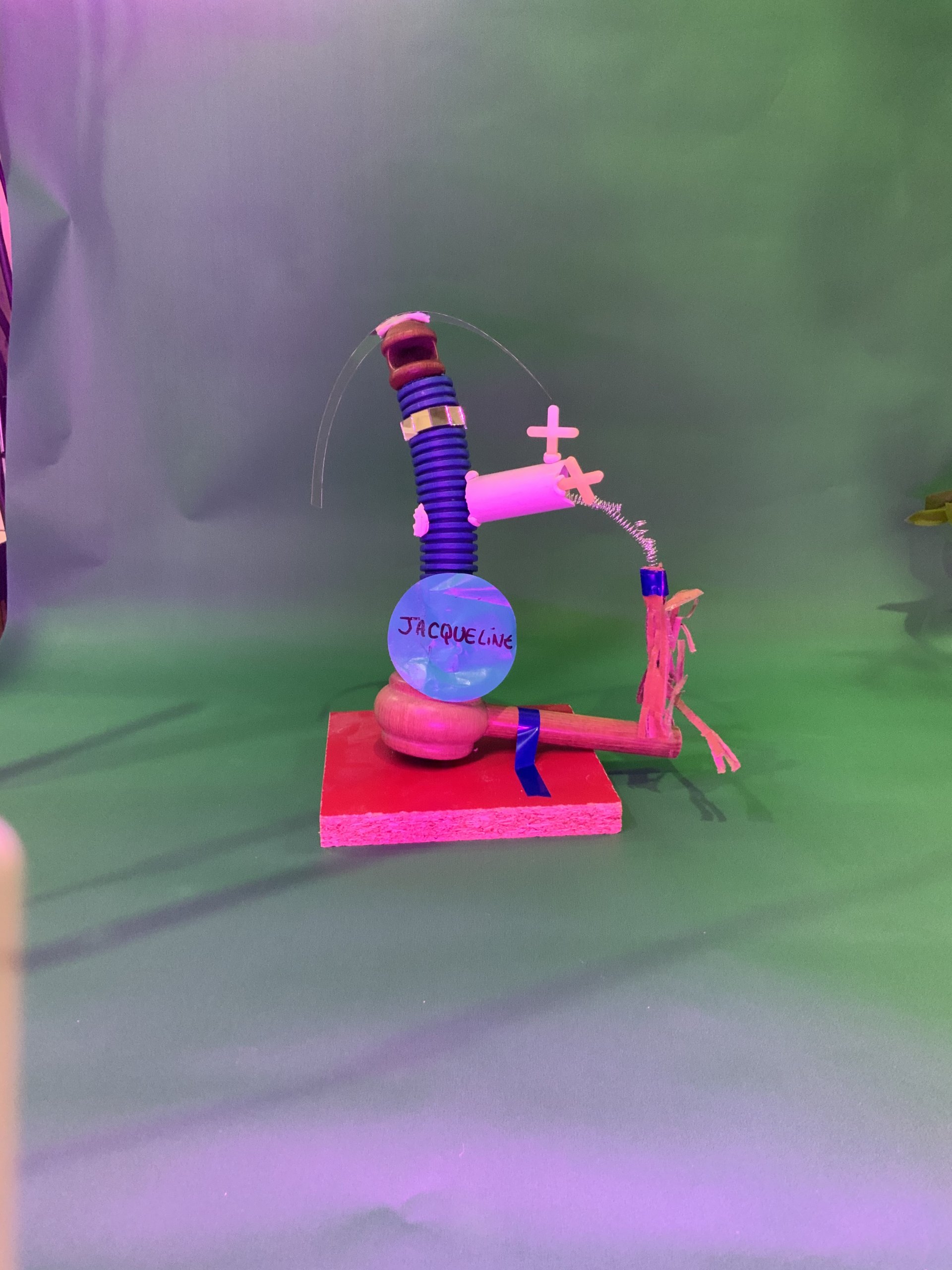 Sculpture mixte intitulée Jacqueline, composée d'un socle bleu en forme de bouteille posé sur une plateforme en bois rouge, sur laquelle sont fixés divers objets, dont une ficelle, un tube en plastique et une croix rose. Le fond est vert.