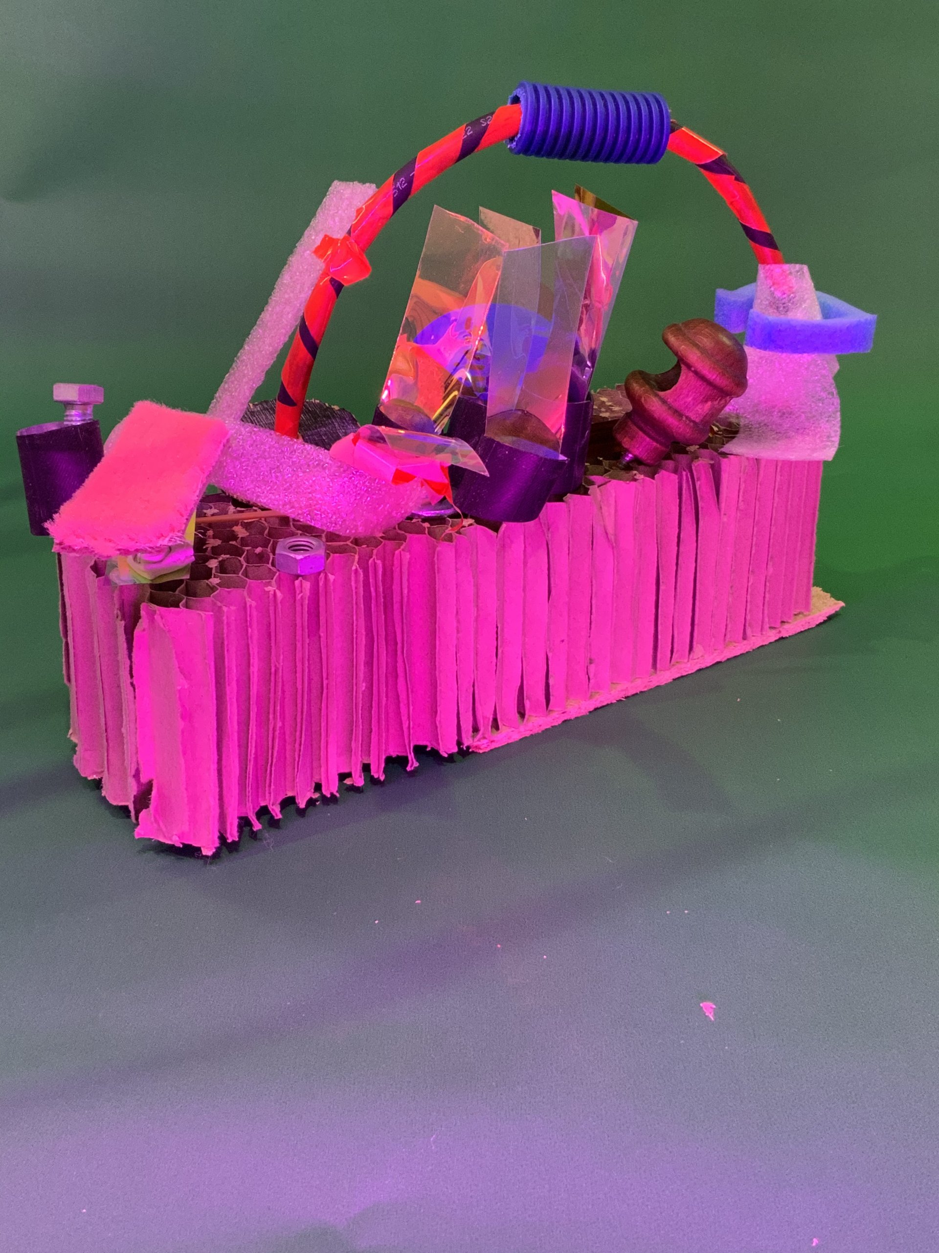 Une sculpture rectangulaire faite de carton ondulé, de pièces métalliques, de mousse, de plastique et d'une poignée rouge et bleue repose sur une surface verte avec un éclairage vert et rose.