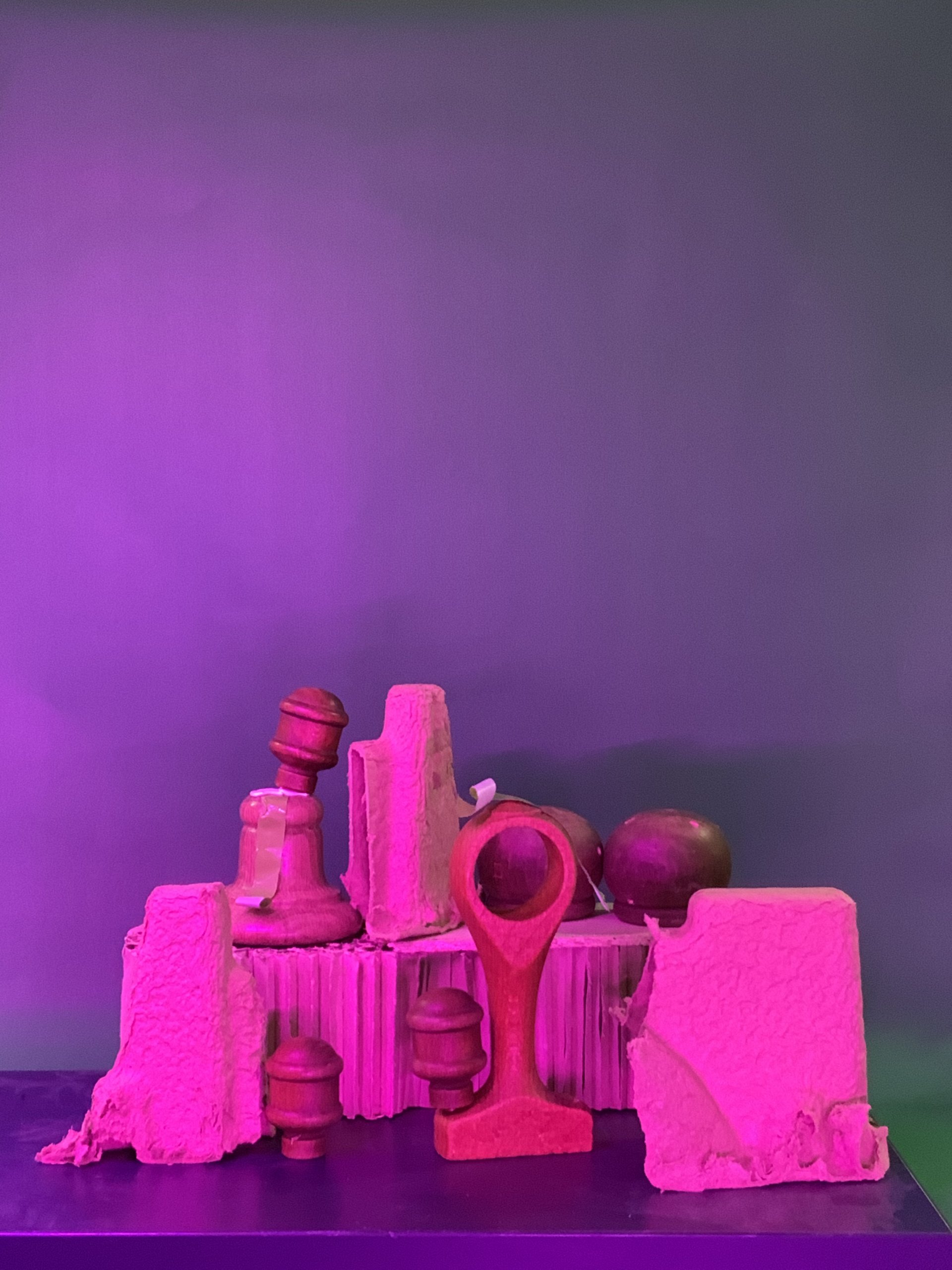 Un groupe d'objets sculpturaux rouges et roses, comprenant des pièces d'échecs et des blocs texturés, disposés sur un fond violet et vert sous un éclairage rose.