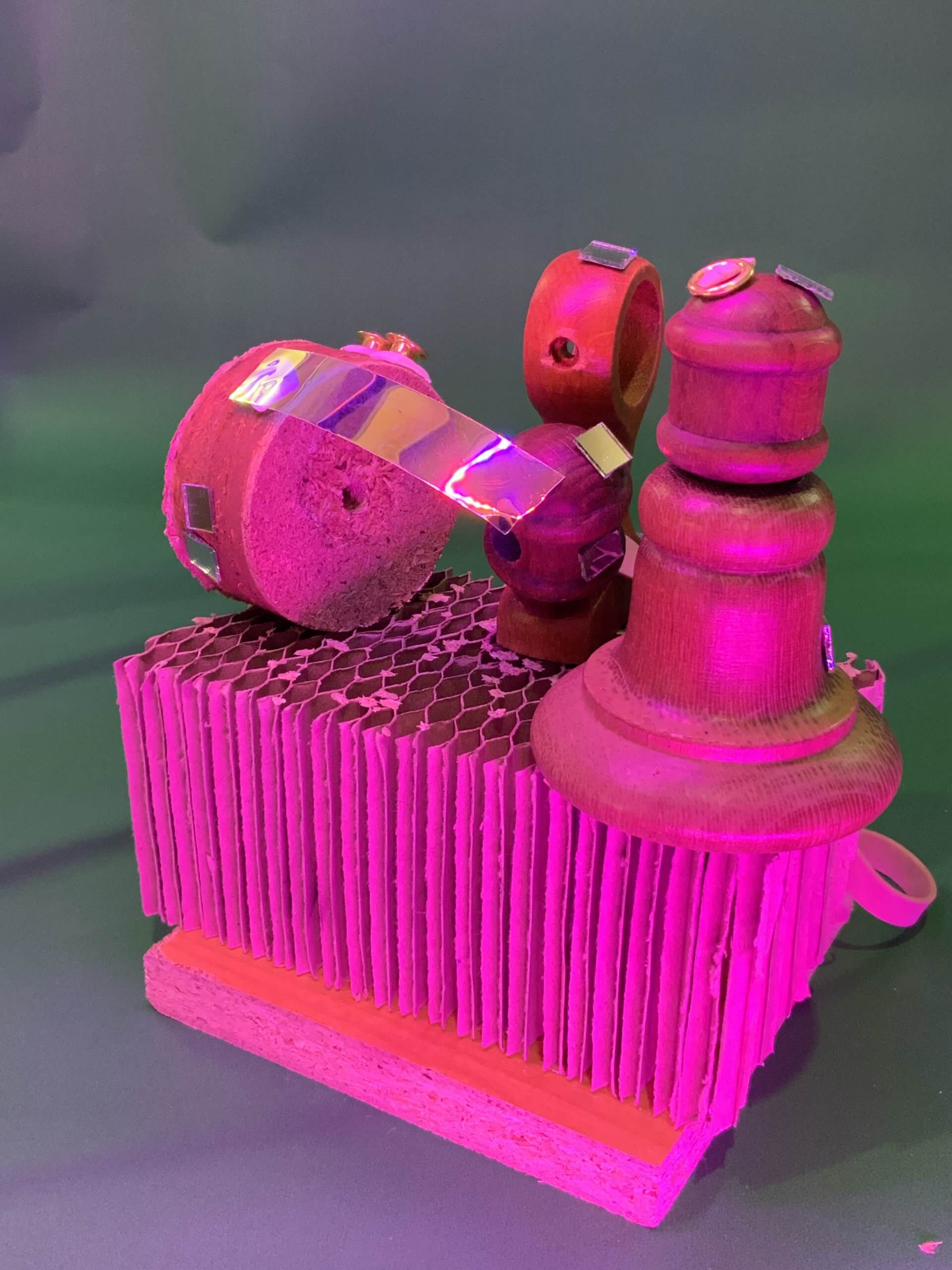 Une pièce d'échecs en bois et un petit objet en bois avec un bouchon en liège et du ruban métallique brillant sont positionnés sur une base en carton texturé, le tout éclairé par un éclairage rose et vert.