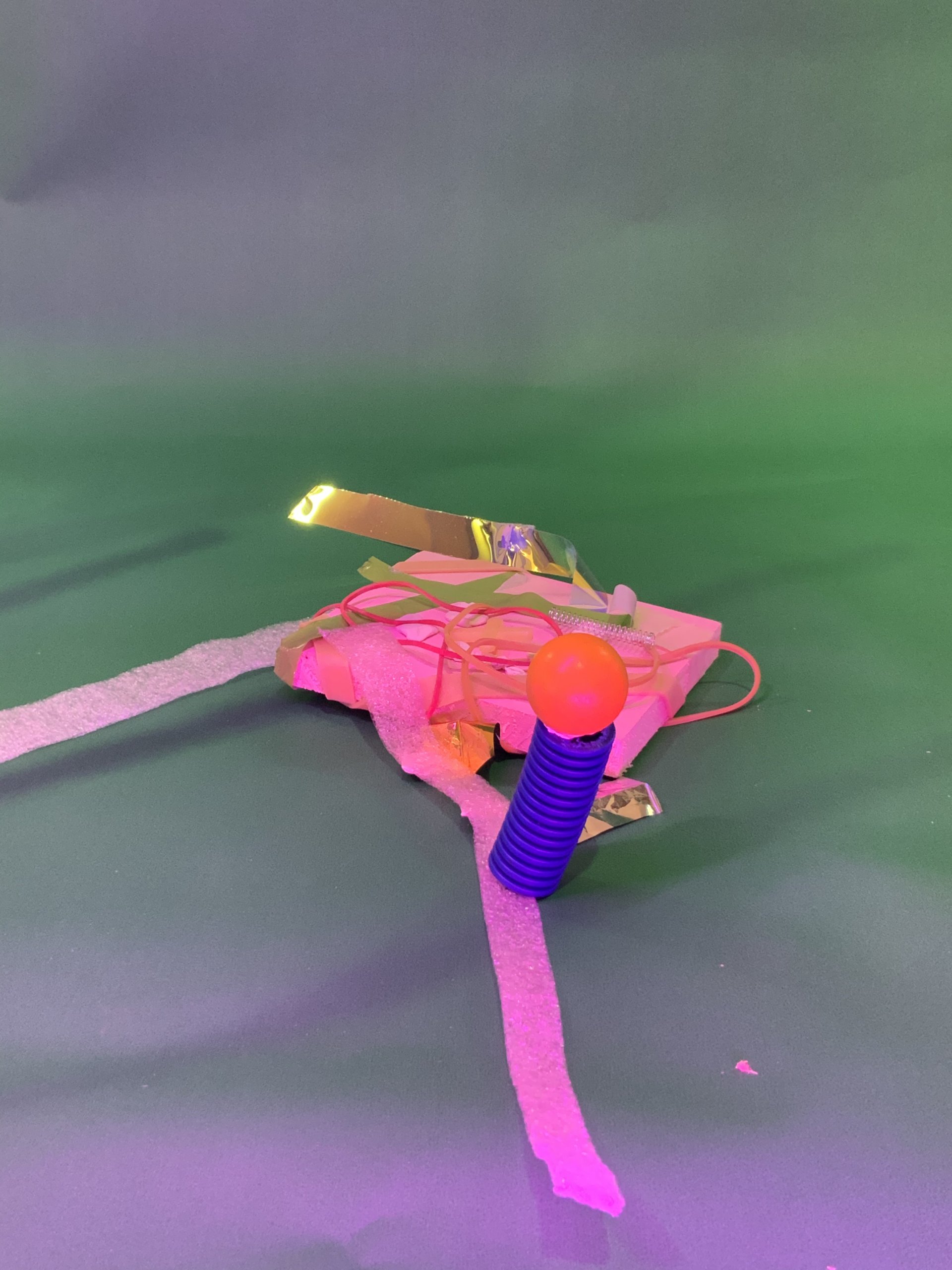 Un cylindre bleu strié avec une boule orange sur le dessus se dresse devant un tas de mousse rose, de feuilles d'or, de fils et de bandes de tissu blanches, le tout sur un fond vert avec un éclairage violet.