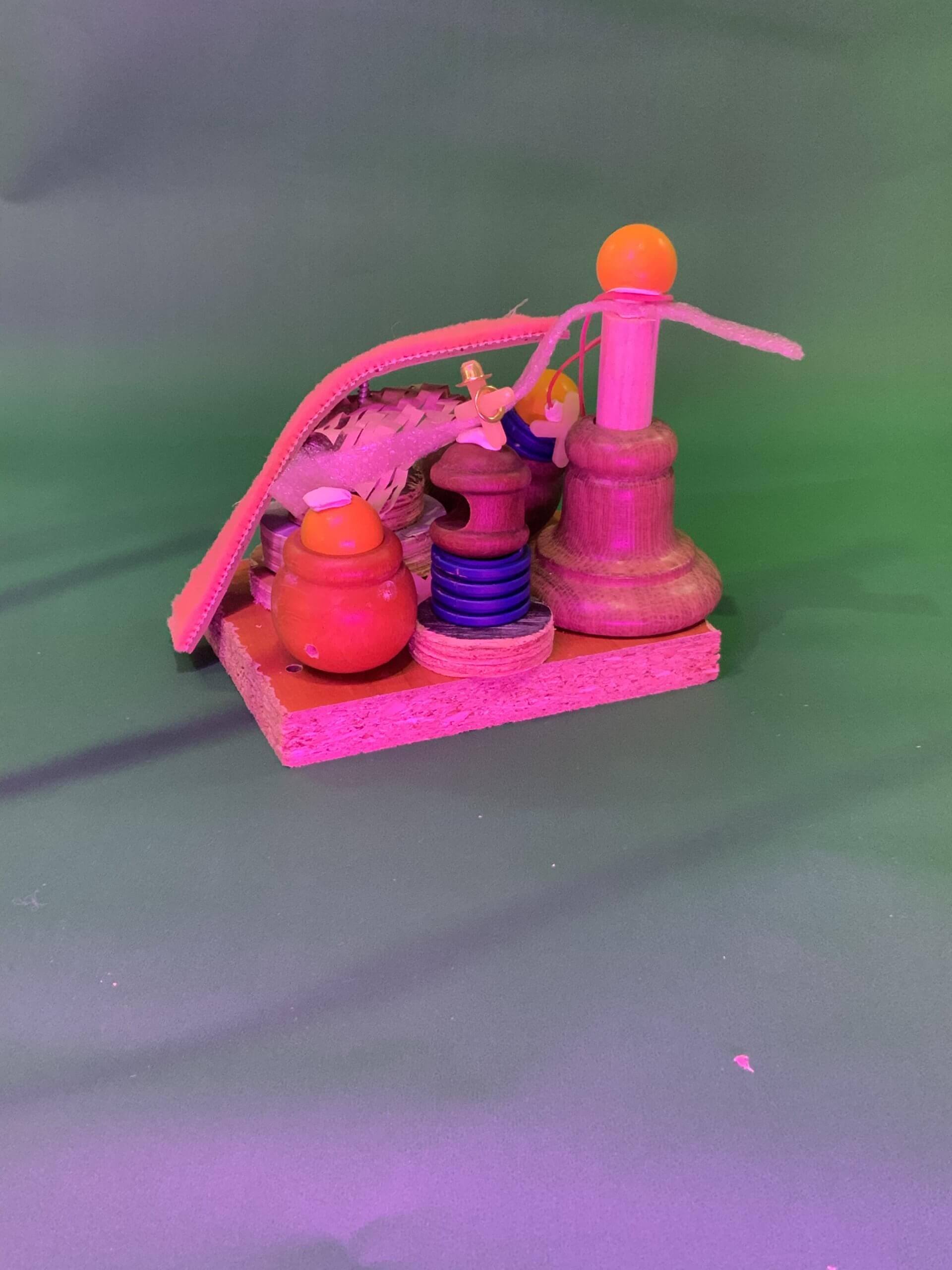 Une petite sculpture abstraite composée d'objets en bois, en métal et en plastique repose sur un socle carré en bois. Elle est composée de bobines, d'un petit pot, d'une plume, d'une paille courbée et d'une boule orange, le tout disposé sur un fond vert.