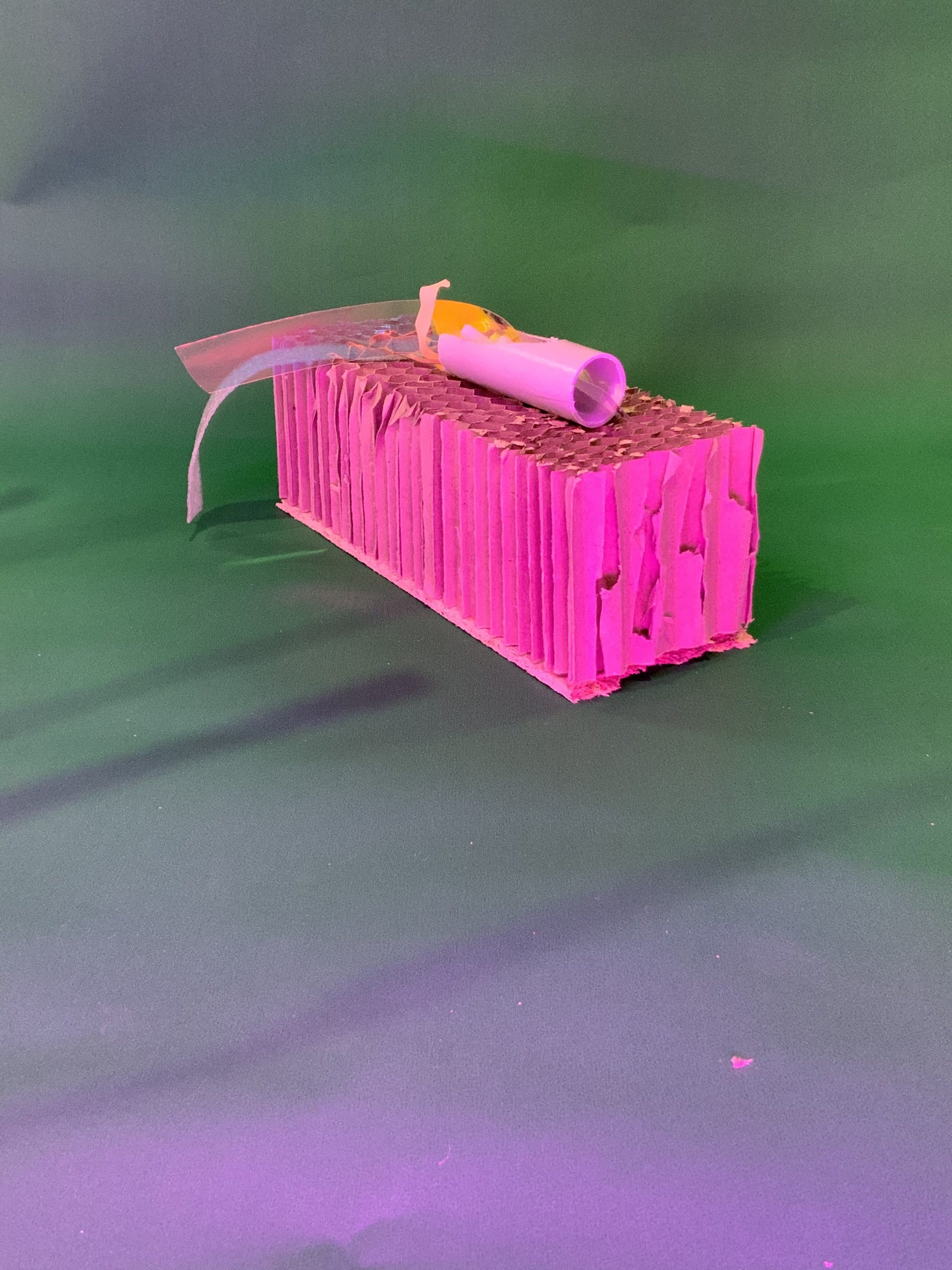 Un bloc rectangulaire en carton ondulé rose repose sur une surface verte avec un fond vert. Un tube de colle jaune et blanche et une bande de ruban adhésif sont placés dessus.