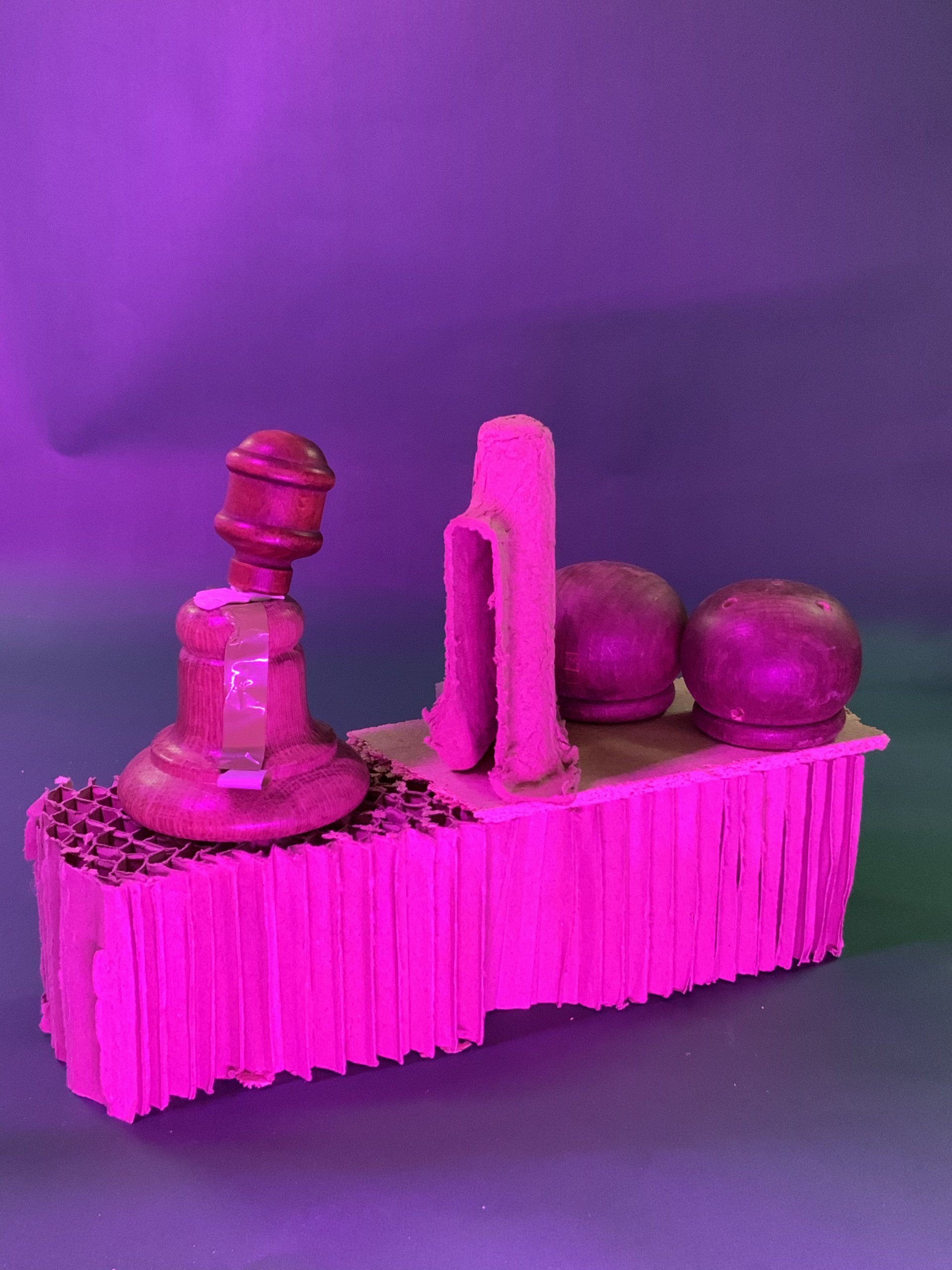 Un arrangement sculptural composé d'objets en bois, dont un marteau et deux sphères, placés sur une base en carton texturé, le tout éclairé par un éclairage rose et violet sur un fond uni.