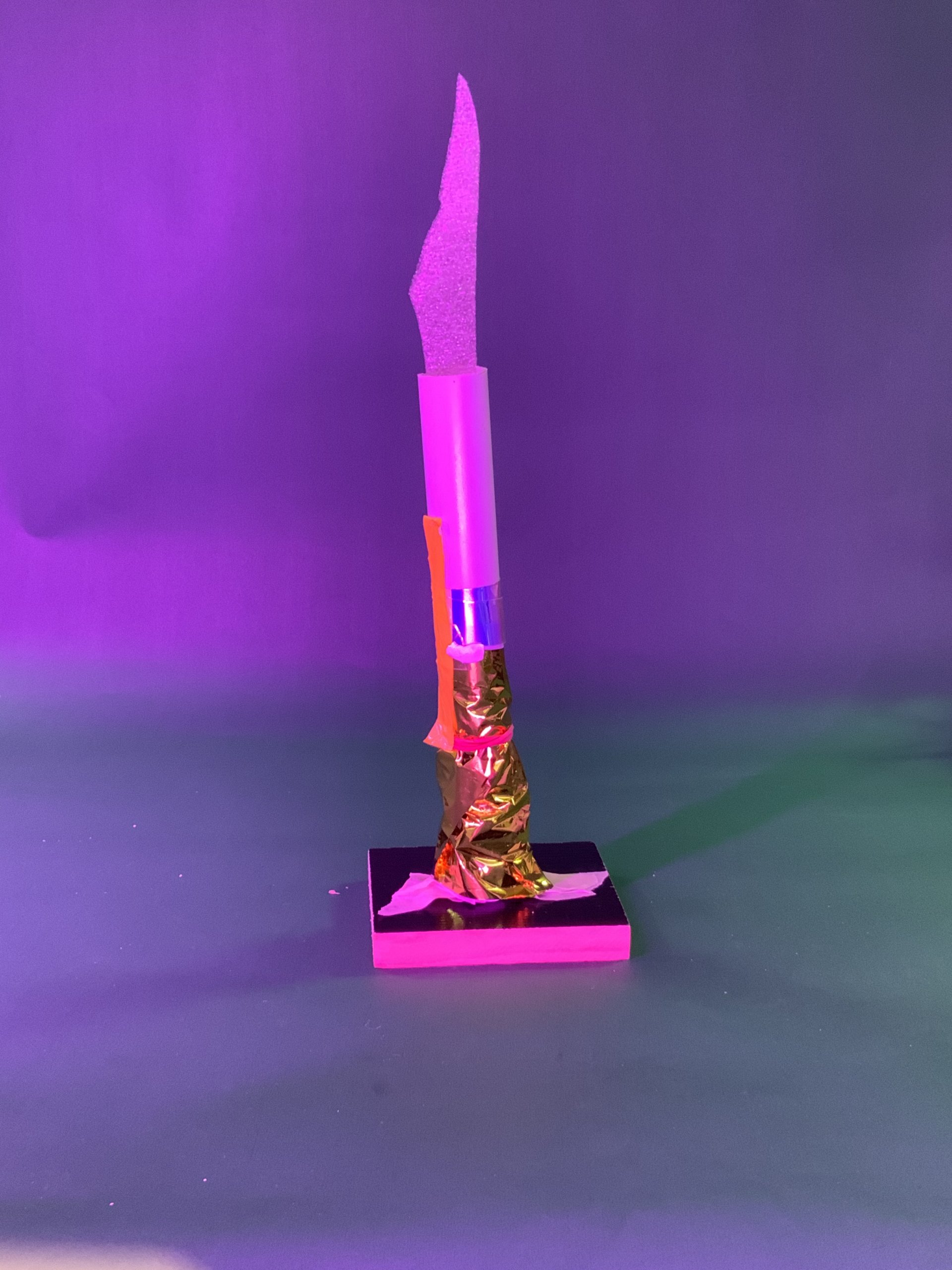 Une sculpture abstraite verticale composée d'un morceau de mousse rose, d'un tube transparent et d'une feuille d'or sur une base rectangulaire noire, sur un fond violet avec un éclairage vert et violet.