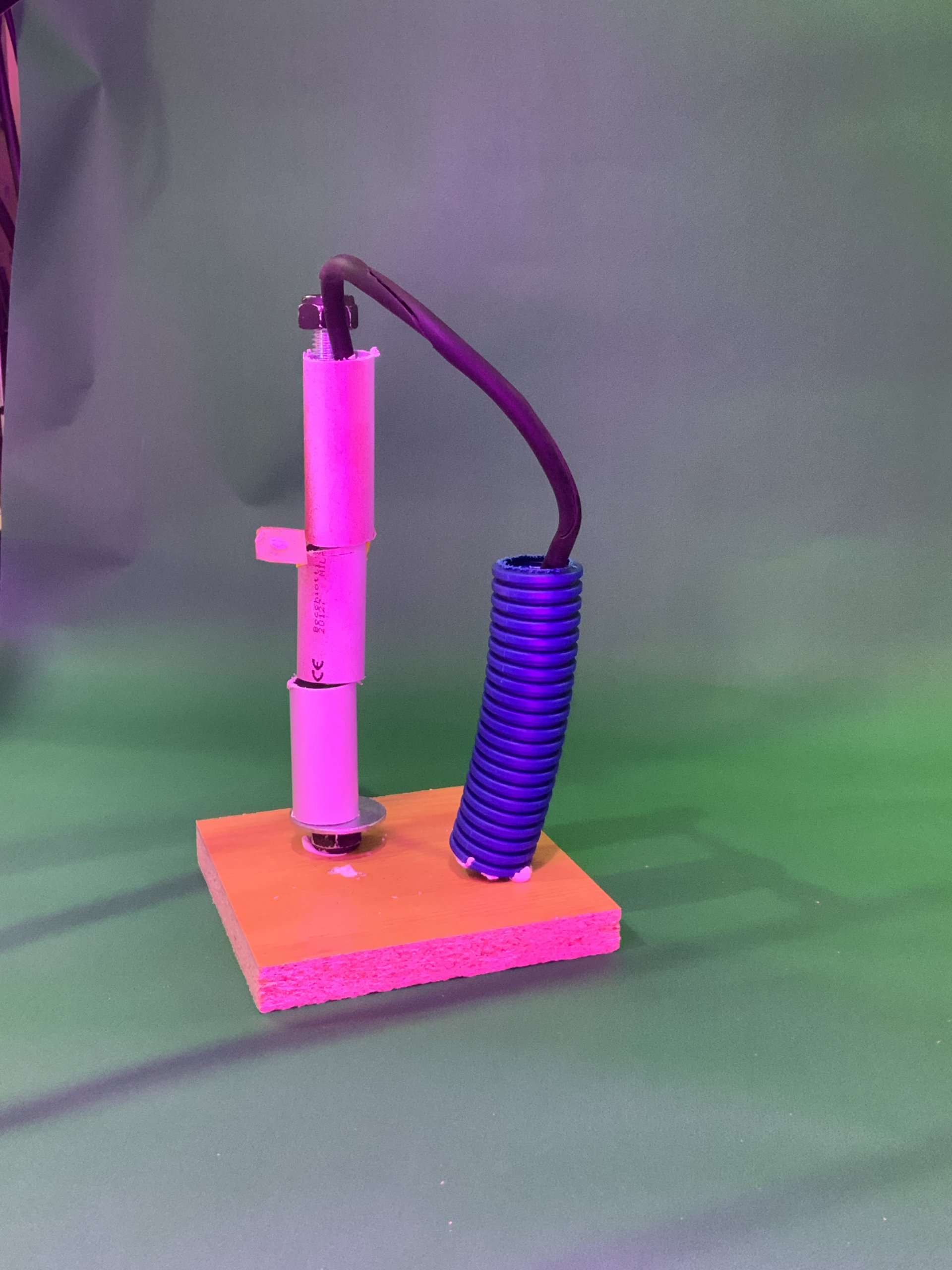 Un dispositif de bobine électrique fait maison monté sur une base en bois, comportant un composant cylindrique blanc vertical et un fil enroulé bleu à côté, sur un fond vert.