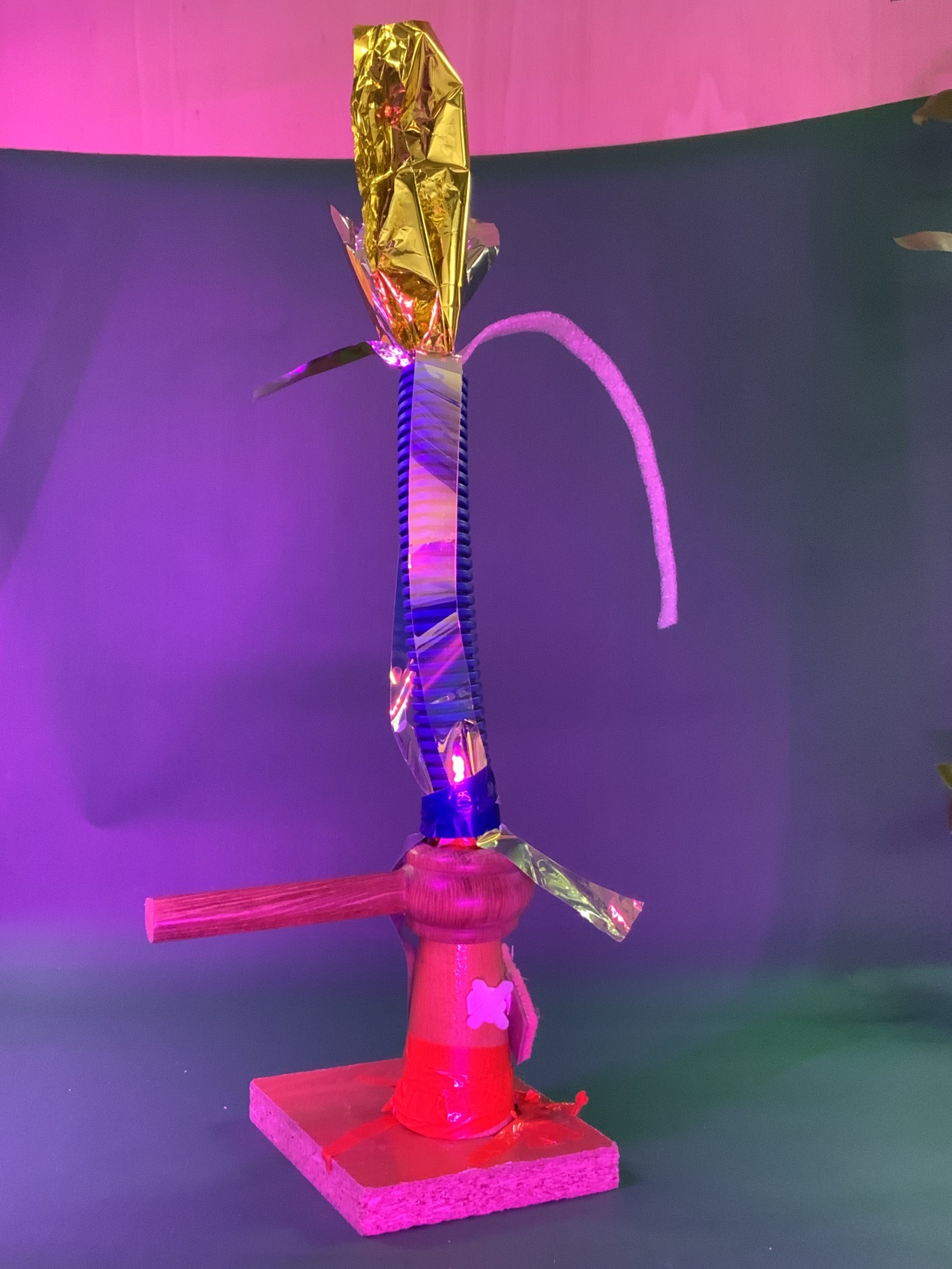 Une sculpture verticale réalisée à partir d'une base en bois, d'une bobine, d'un cylindre rouge, d'un bâton, d'un ruban bleu, d'un cure-pipe blanc et d'une feuille d'or, sur un fond vert avec un éclairage rose et violet.