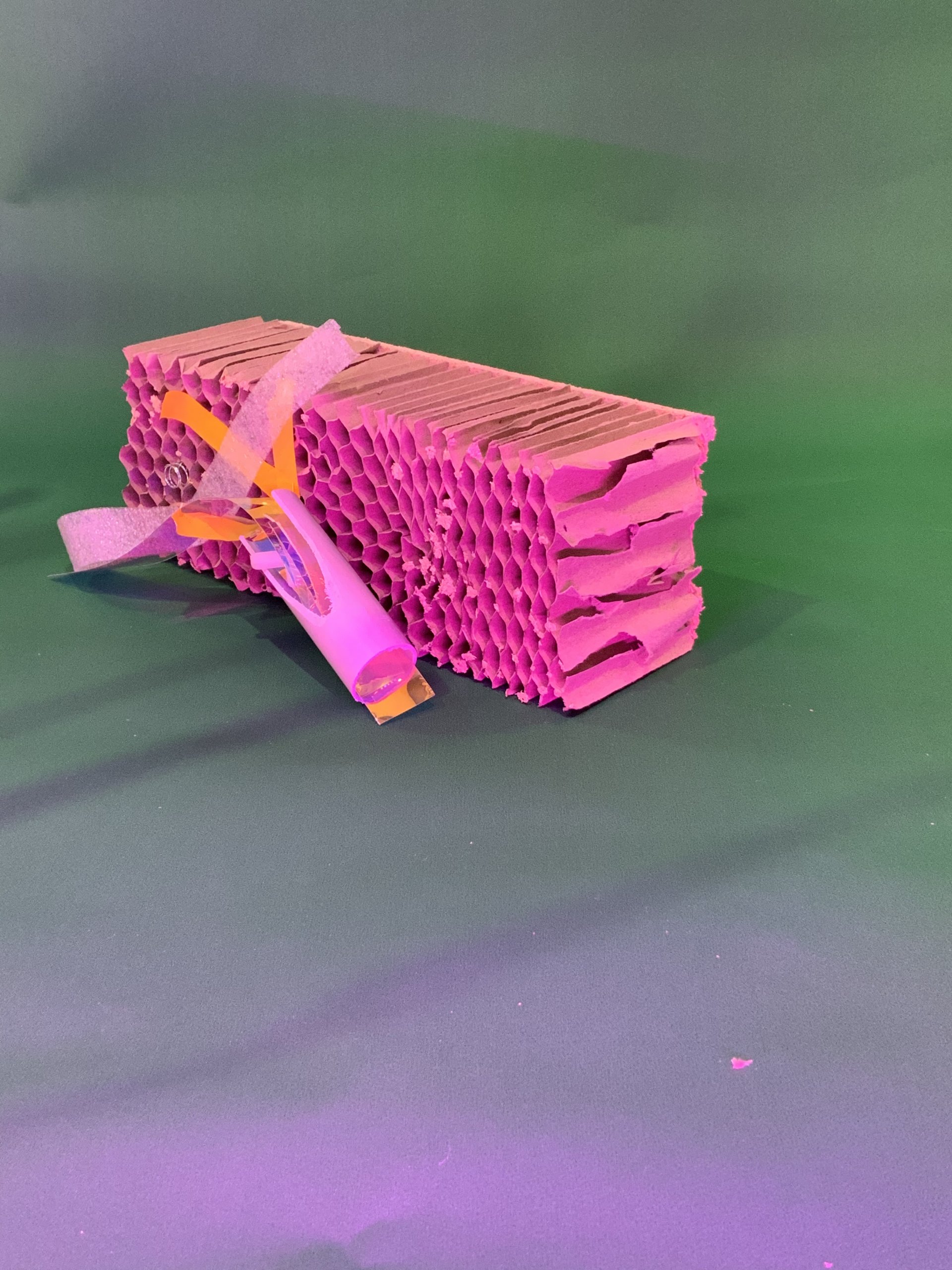 Une pile rectangulaire de carton alvéolé rose repose sur un fond vert. Devant elle se trouve un petit objet cylindrique enveloppé de papier violet orné de bandes de papier orange et jaune.