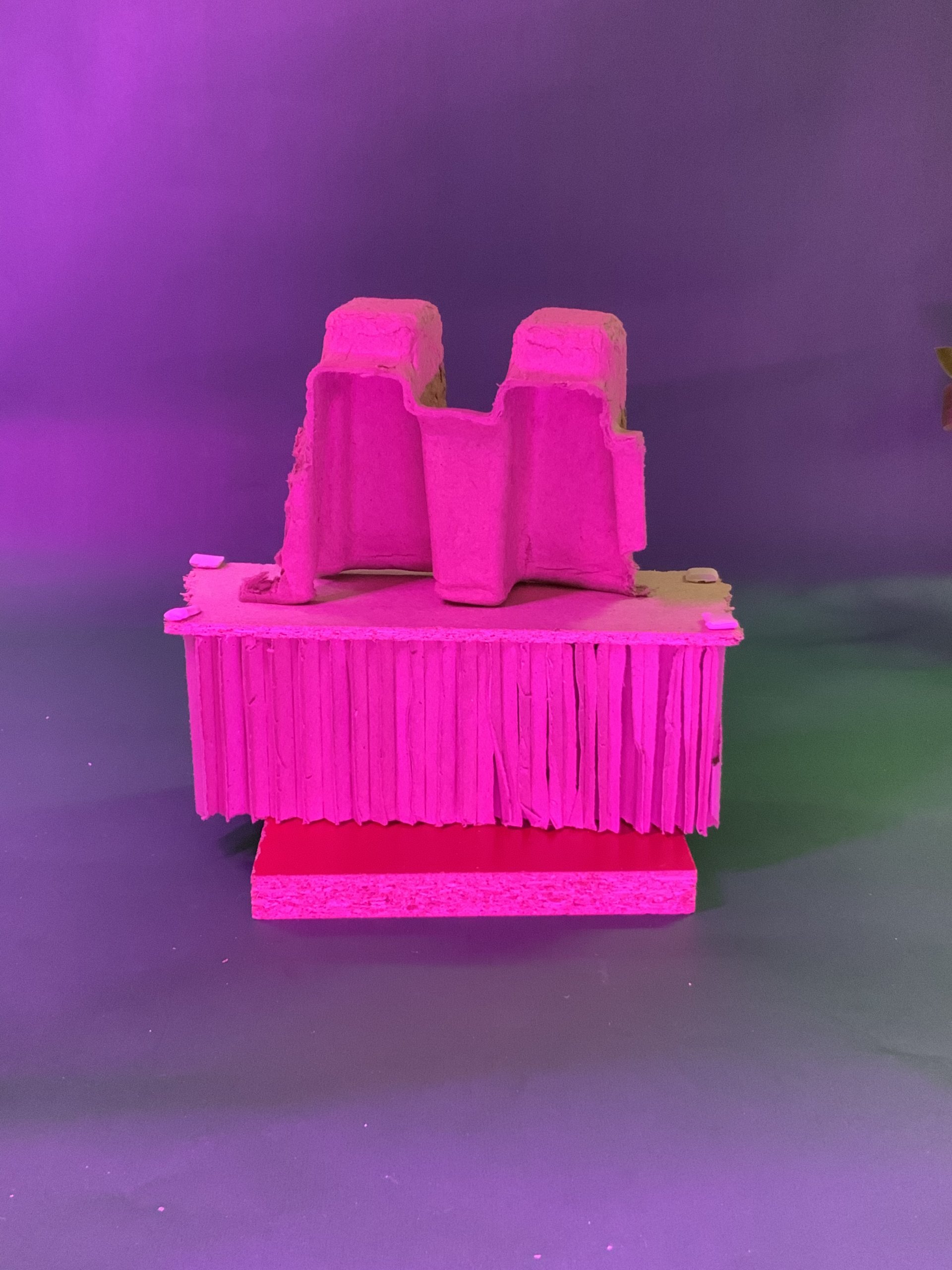Un arrangement sculptural de matériaux en carton et en papier empilés sur une plate-forme rose, avec du papier à franges au milieu et une pièce ondulée sur le dessus, le tout éclairé par un éclairage rose et violet.