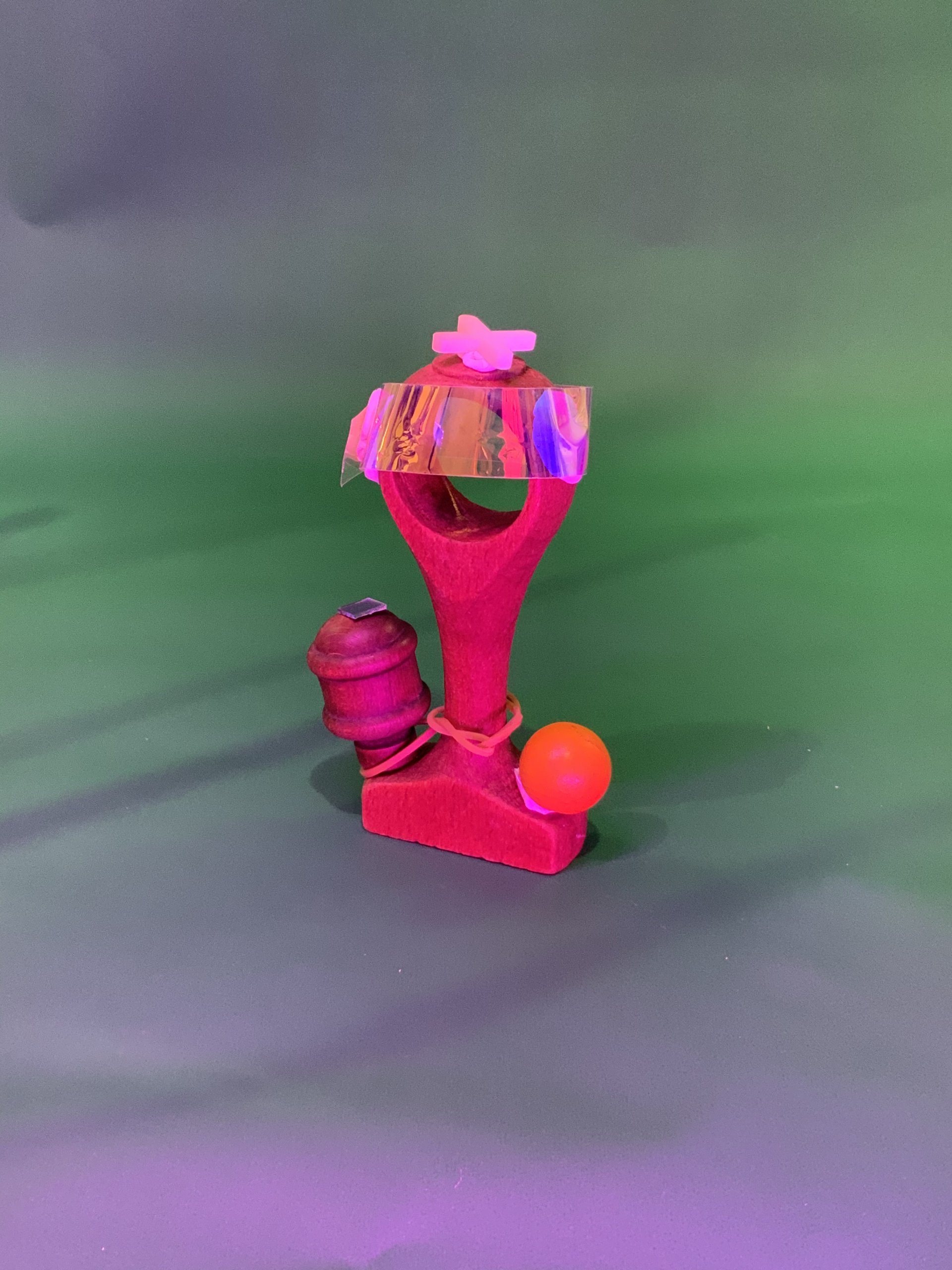 Une petite sculpture de fronde faite à la main avec une base rose, une pièce en miroir attachée sur le dessus, un élastique, une balle orange et un objet cylindrique sur le côté, sur un fond vert.