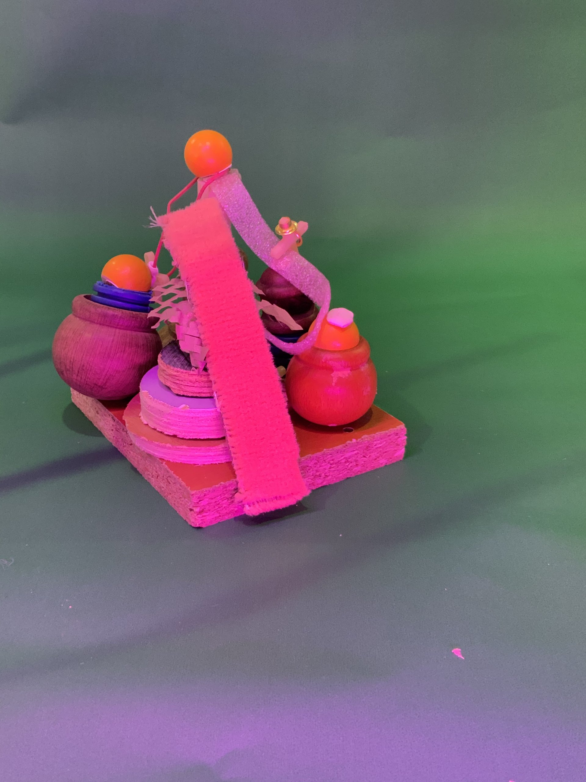 Une petite sculpture abstraite composée d'objets en bois, de disques colorés, d'une bande texturée rose vif et de boules orange, le tout disposé sur une base carrée en bois sur un fond vert avec un éclairage rose.