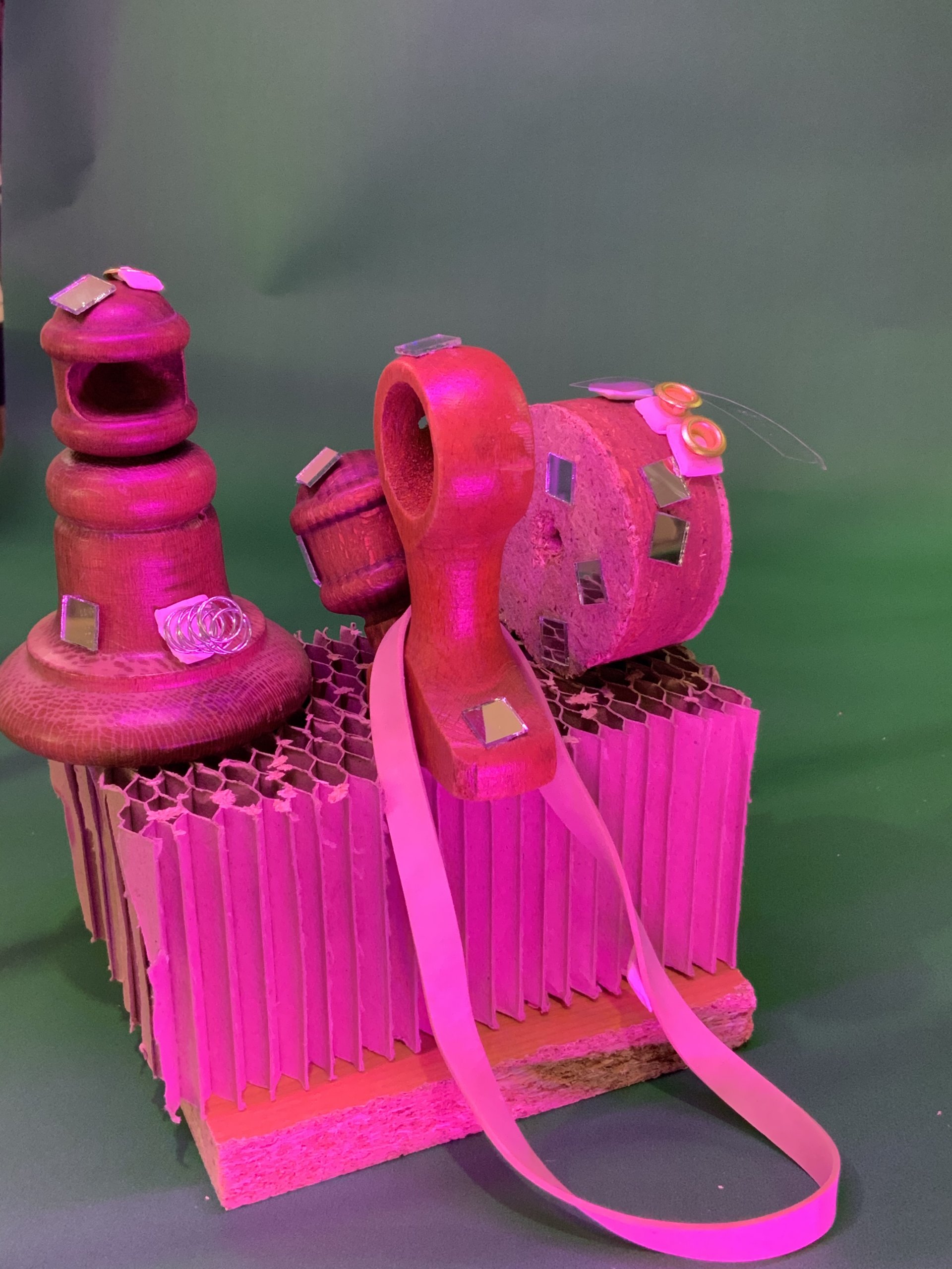 Un ensemble d'objets en bois, ornés de miroirs et de touches de rose, est disposé sur un socle en carton ondulé sur fond vert, sous un éclairage rose. Un ruban rose recouvre l'ensemble.