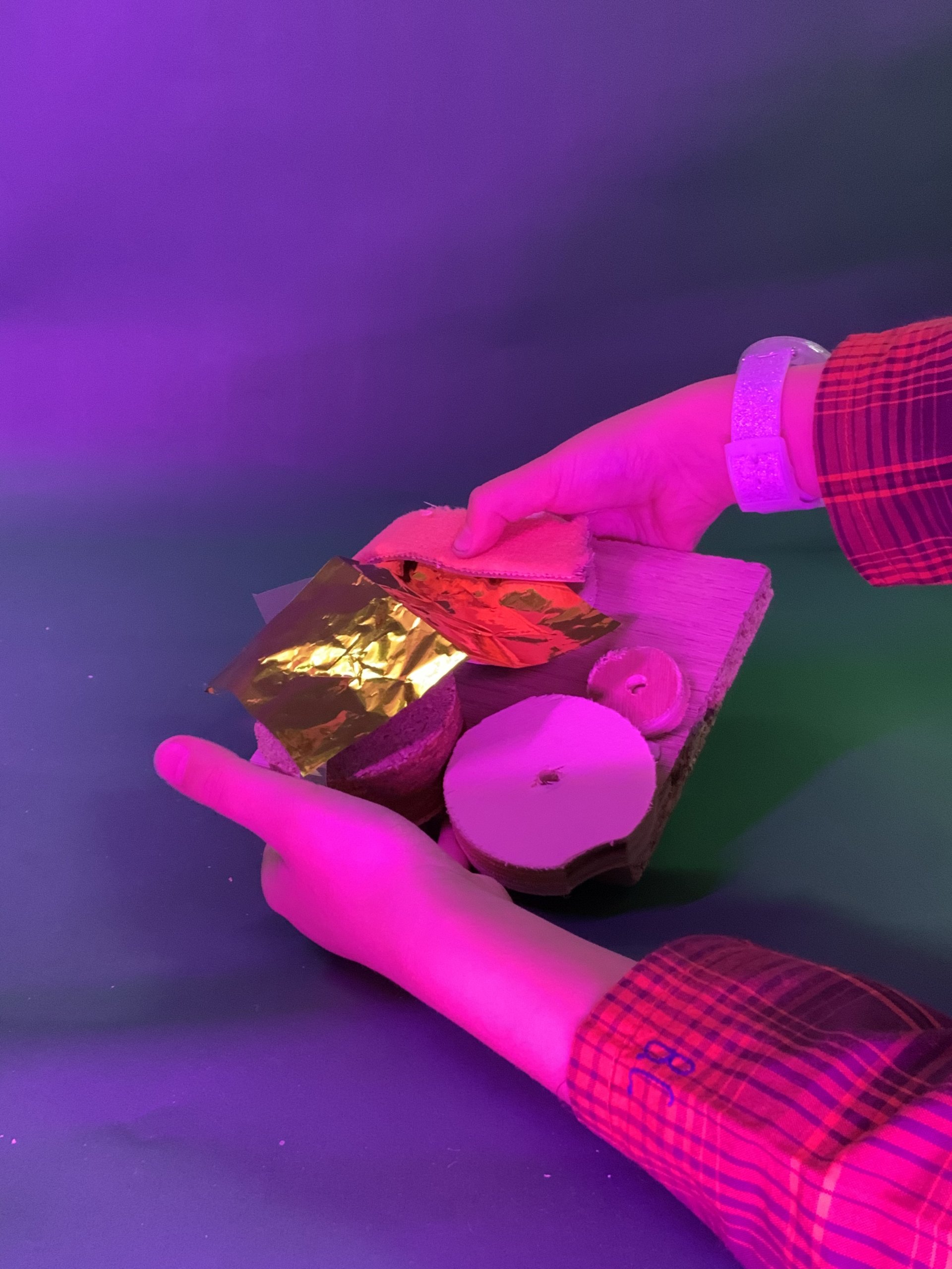 Deux mains tiennent une petite variété de matériaux, notamment des tranches de bois, un bouchon en liège et un morceau de feuille d'aluminium de couleur dorée, sous un éclairage rose et violet sur un fond vert et violet.
